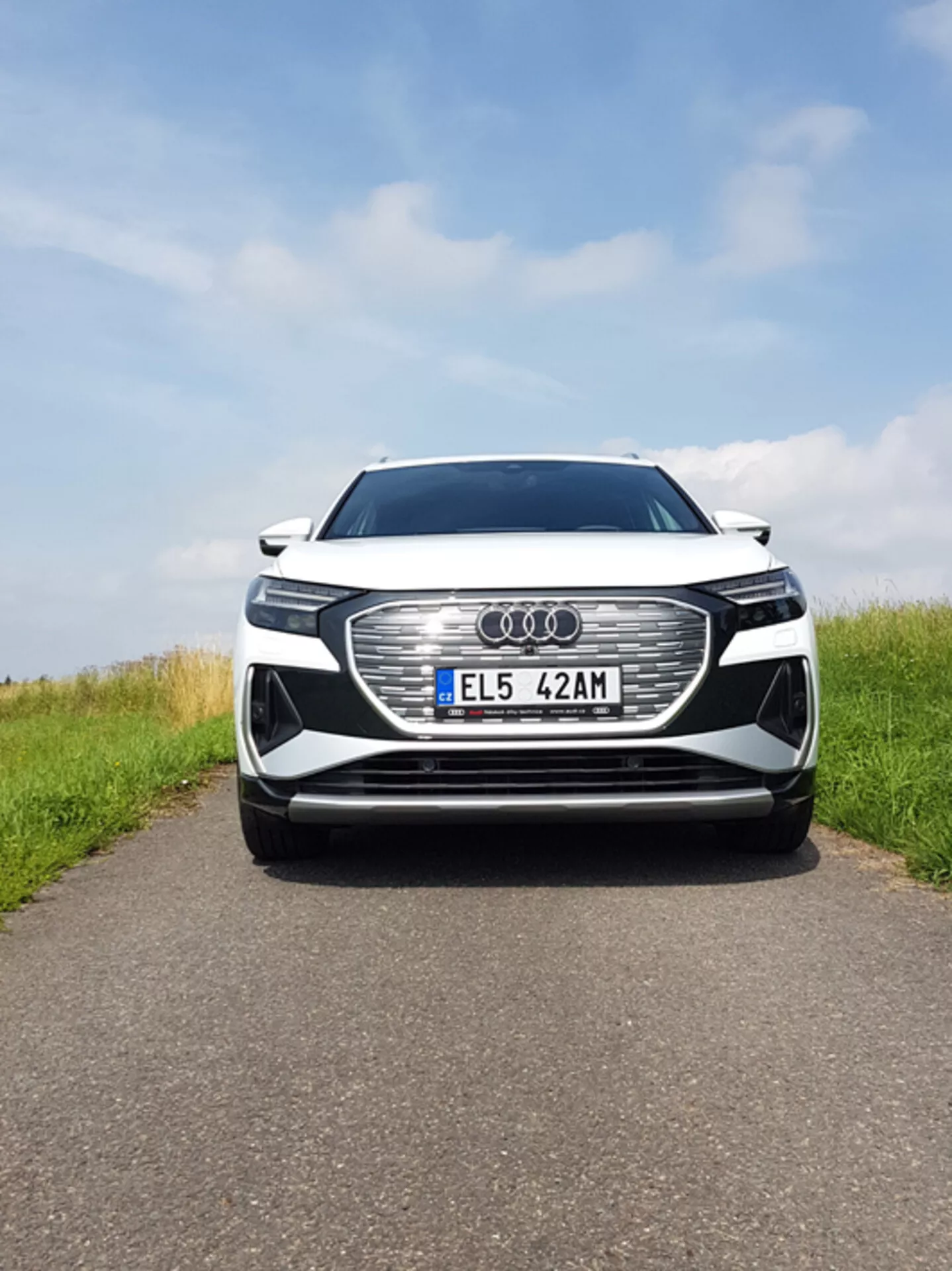 Wyśrodkowany przedni widok Audi Q4 e-tron 2021, przedstawiający dużą heksagonalną kratę z emblematem Audi, światła LED, europejską tablicę rejestracyjną, potencjalne sensory ADAS, wloty powietrza, metaliczny lakier i detale chromowane pod częściowo zachmurzonym niebem na asfaltowej drodze z trawiastymi bokami.