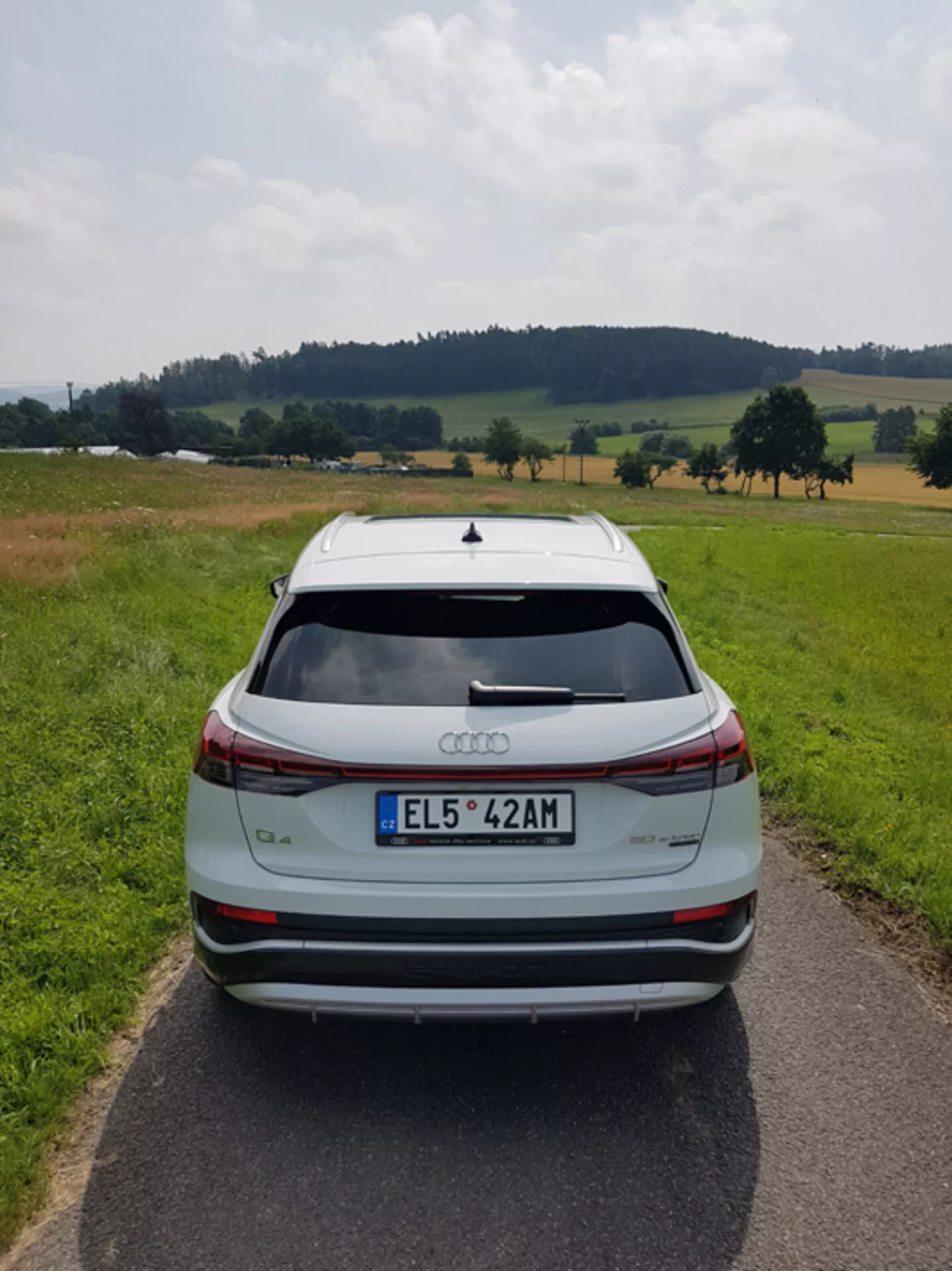 Tylny widok srebrnego Audi Q4 e-tron 2021 na wąskiej brukowanej drodze pośród pasterskiego krajobrazu z polami i drzewami pod częściowo zachmurzonym niebem, podkreślający jego elegancki design, świetlne belki hamulcowe, spoilera dachowego, czujniki parkowania z tyłu i dwukolorowe obręcze aluminiowe.