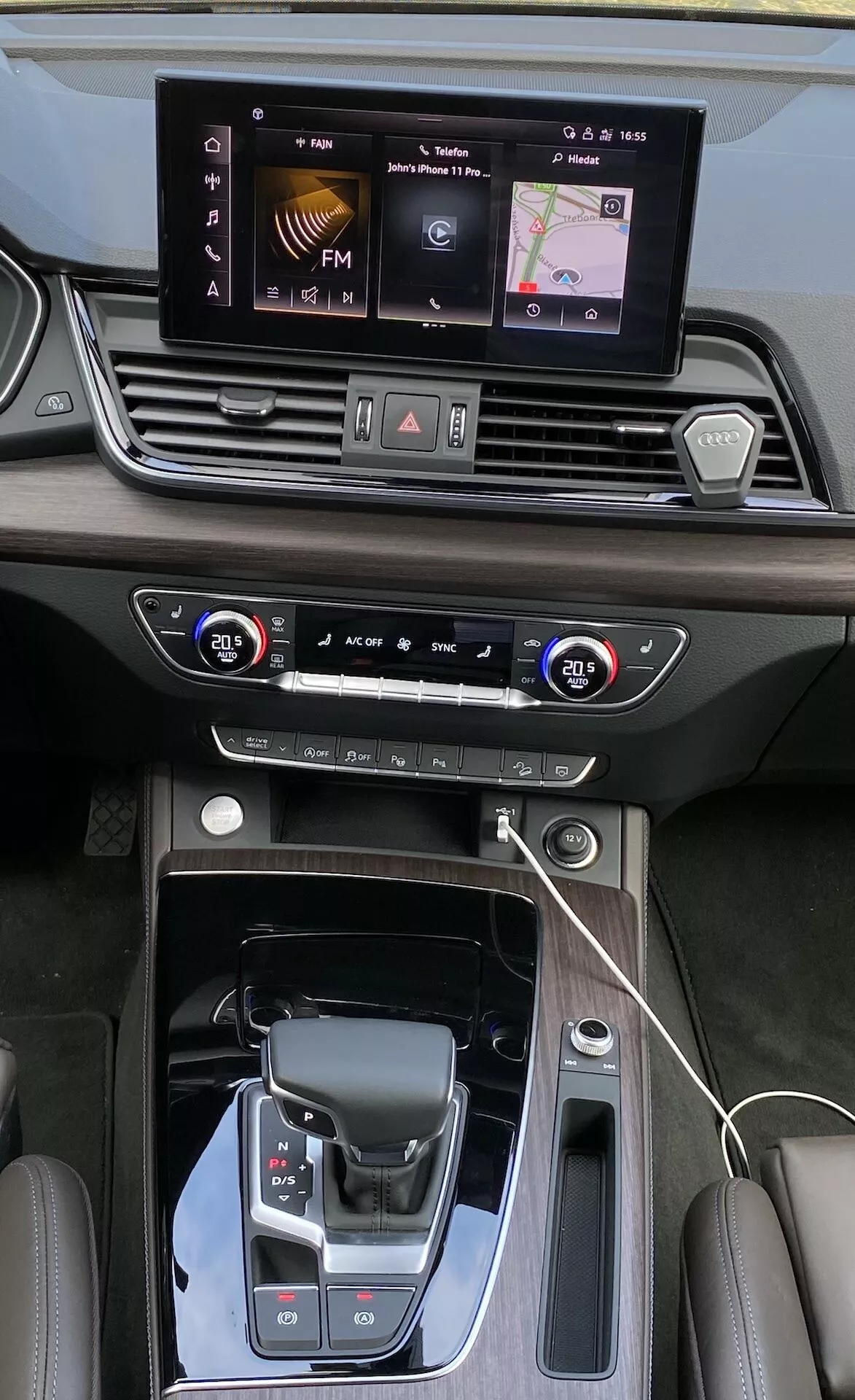 Widok z góry na konsolę środkową i deskę rozdzielczą Audi Q5 2020 Sportback, pokazujący szczegółowe cechy, takie jak dotykowy wyświetlacz infotainment, sterowanie klimatyzacją dwustrefową, nowoczesny selektor biegów z osłoną skórzaną i luksusowymi przeszyciami. Perspektywa podkreśla elegancki design, akcenty w fortepianowej czerni i metalu oraz zaawansowane technologie, w tym bezprzewodową ładowarkę i wybór trybu jazdy.