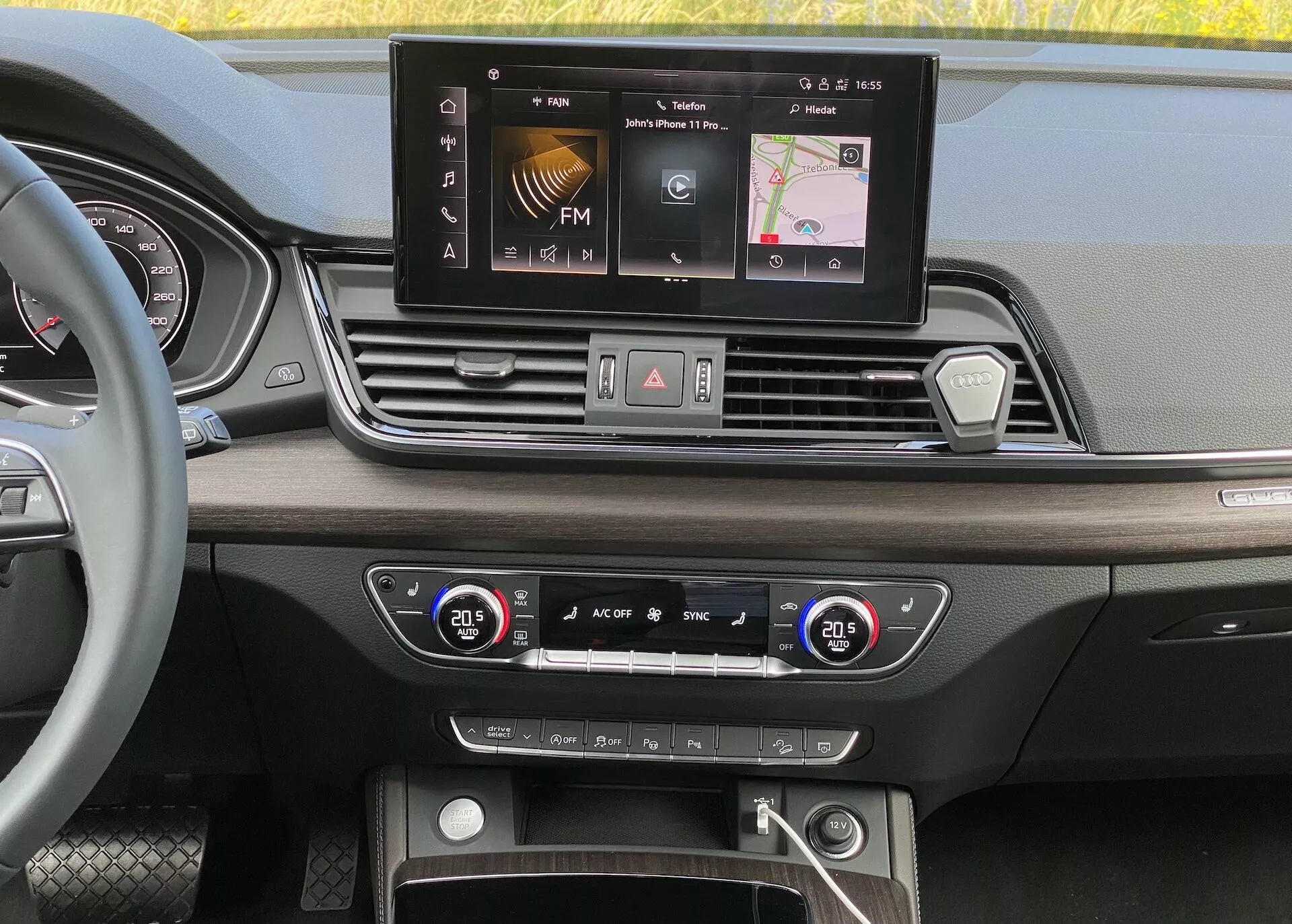 Widok wnętrza deski rozdzielczej Audi Q5 2020 Sportback z perspektywy poziomu oczu. Obrazek podkreśla szerokoekranowy wyświetlacz infotainment z opcjami takimi jak radio FM, nawigacja i ustawienia pojazdu. Poniżej widoczne są cztery symetrycznie rozmieszczone kratki wentylacyjne, centralna konfiguracja przycisków i gałek do świateł awaryjnych, odszraniania i szybkiego dostępu do ustawień. System dwustrefowej klimatyzacji z cyfrowymi wyświetlaczami temperatury jest ustawiony na 20,5 stopnia Celsjusza dla kierowcy i pasażera. Dolna część ukazuje schowek z możliwymi portami USB, wejściem pomocniczym i przyciskiem start/stop do technologii bezkluczykowego zapłonu. Luksusowe materiały i design obejmują wykończenia drewnopodobne i wypolerowane metalowe akcenty.