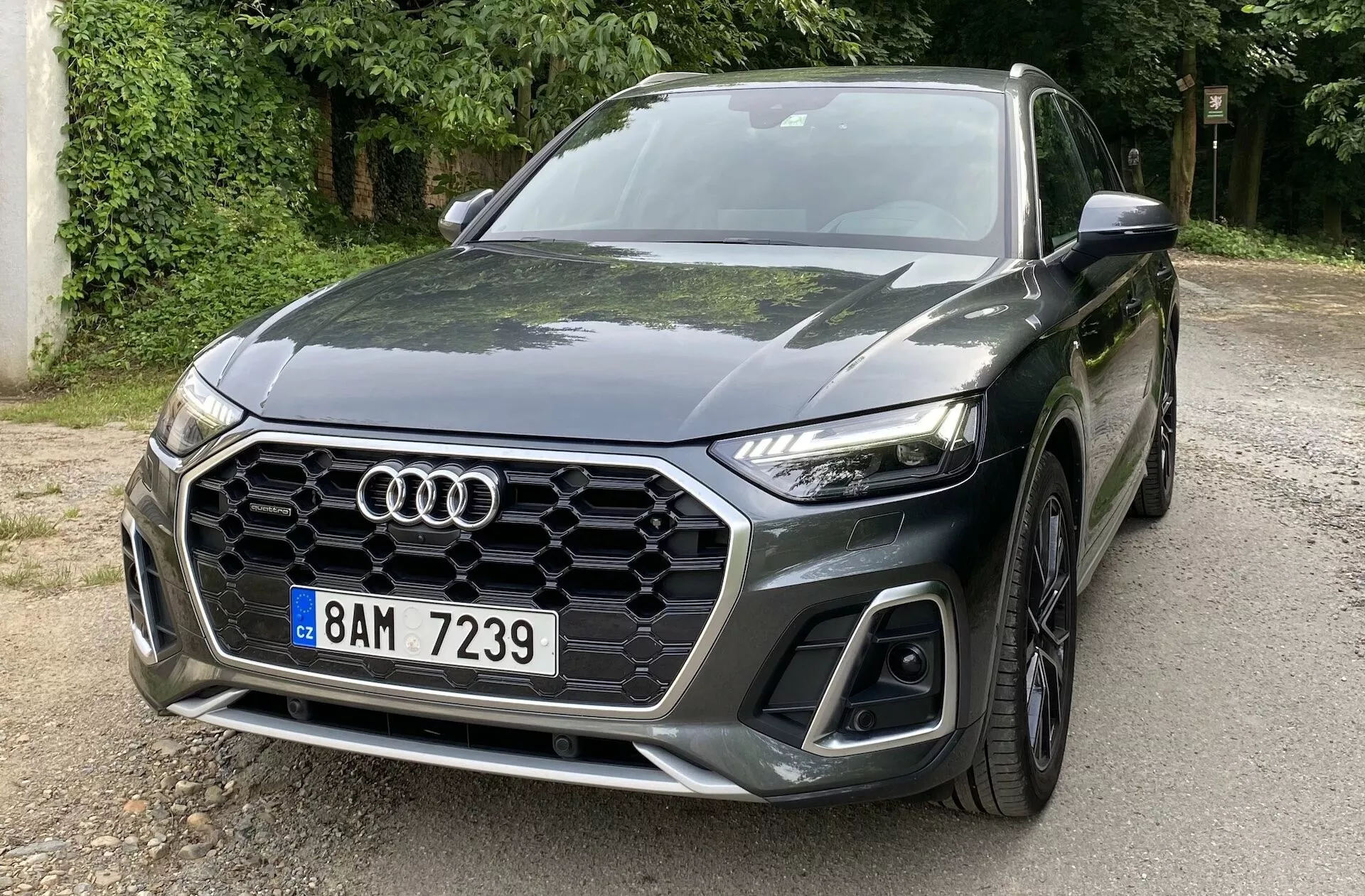 Widok z przodu w trzech czwartych Audi Q5 Sportback 2020, ukazujący przednią, lewą stronę z charakterystycznym, sześciokątnym grillem w kształcie plastra miodu, logo Audi, eleganckimi, kątowymi reflektorami LED i dużymi wlotami powietrza na dolnym zderzaku. Samochód jest zaparkowany na drodze otoczonej drzewami, co odbija się na jego ciemnym, błyszczącym lakierze. Rzeźbiona maska z aerodynamicznymi liniami w kierunku emblematu oraz przednie koło ze stopów z wieloszprychowym wzorem podkreślają jego sportowy i nowoczesny design.
