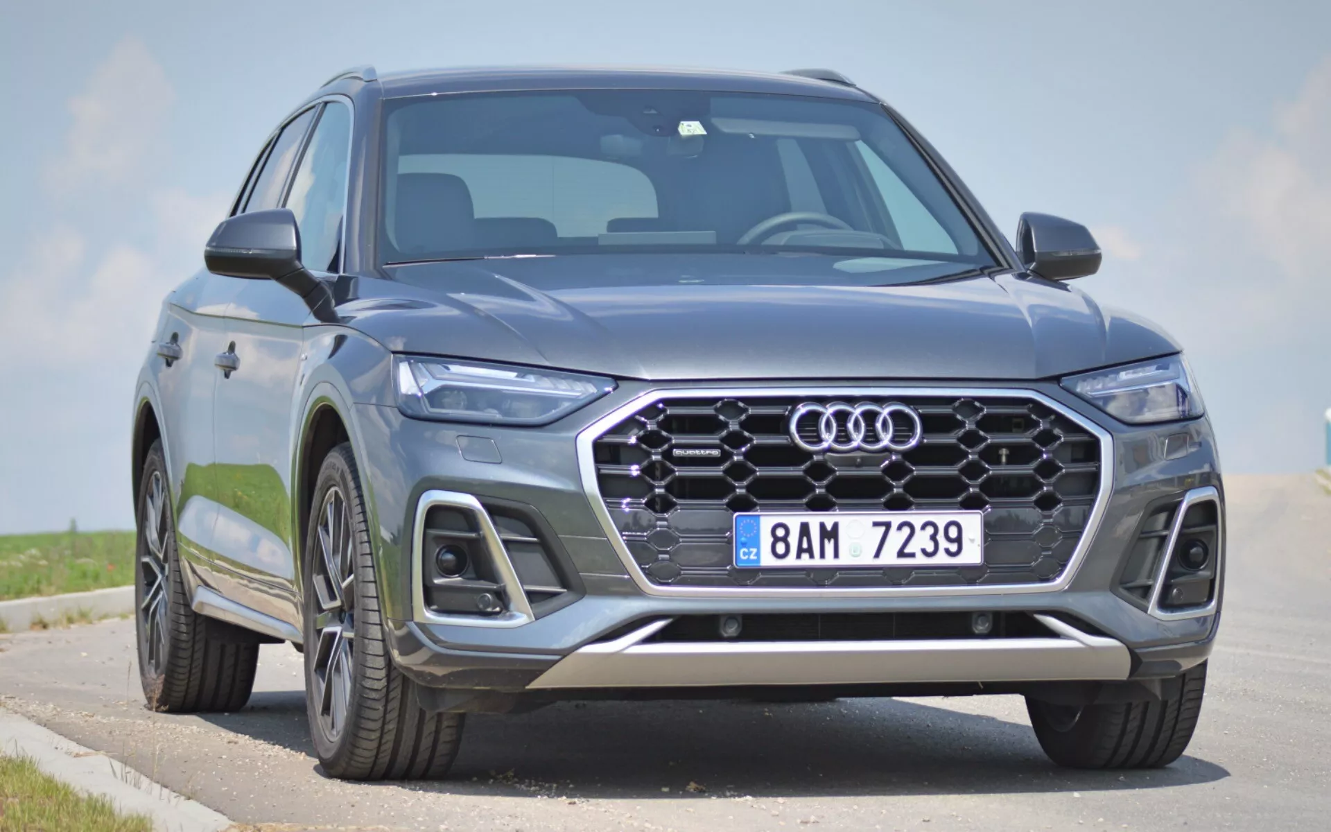 Nowoczesny SUV Audi Q5 2020 (Q5 Sportback) uchwycony z niskiego, przedniego trzech czwartych kąta, ukazujący przedni pas, sześciokątną kratkę z emblematem czterech pierścieni, eleganckie reflektory LED i część bocznego profilu. Duża, wzorzysta kratka, zintegrowane światła przeciwmgielne, wloty powietrza i srebrne elementy wykończeniowe na przednim zderzaku dodają estetyki i funkcjonalności. Zaawansowane technologie oświetleniowe i systemy wspomagania kierowcy sugerowane są przez ostre wzory reflektorów i konfigurację kratki. Duże aluminiowe koła i dynamiczne linie nadwozia wskazują na premium, solidną obecność. Zaparkowany na drodze z czystym niebem w tle, przednie cechy i agresywne stylizacyjne wskazówki pojazdu są podkreślone.