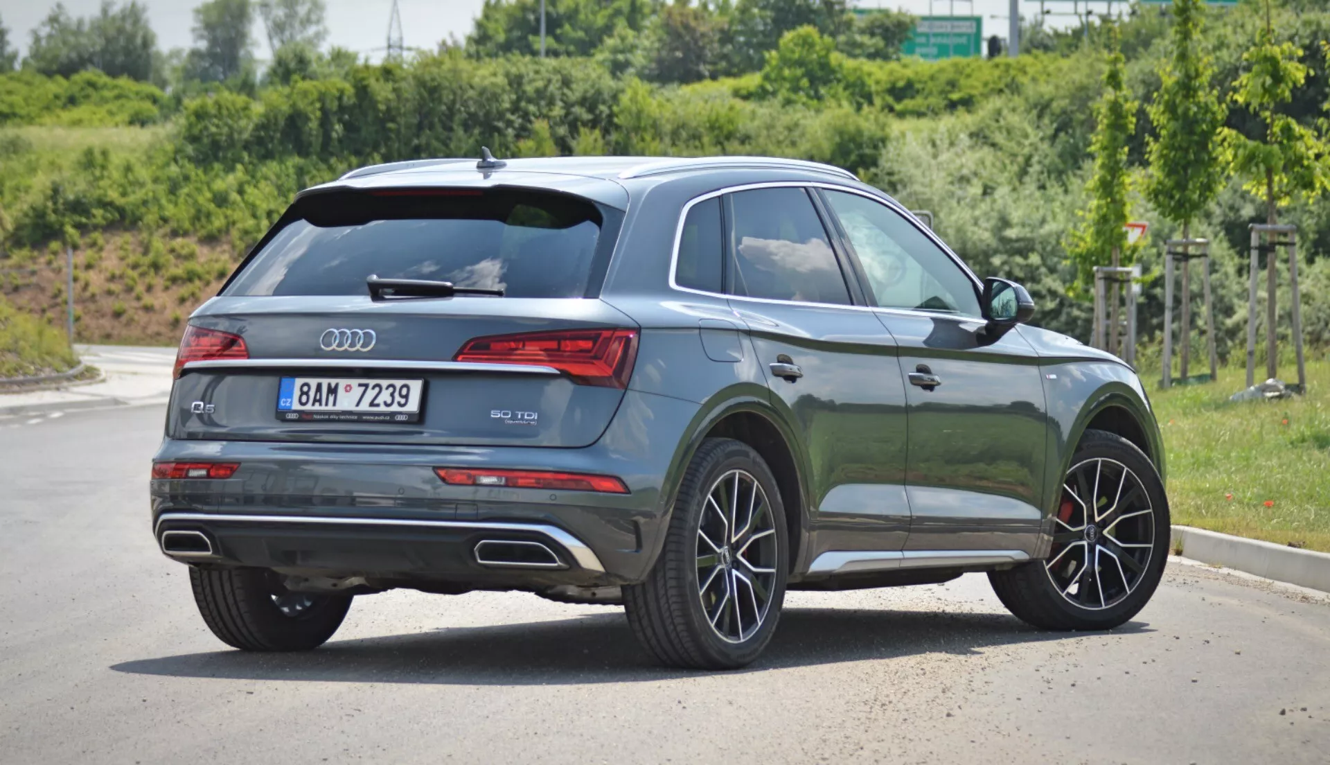 Obraz przedstawia nowoczesne Audi Q5 2020 Sportback SUV zaparkowane na brukowanej powierzchni w ciągu dnia, widziane pod kątem trzech czwartych od tyłu. Tylna klapa zawiera logo Audi i oznaczenie modelu, z eleganckimi, poziomymi tylnymi światłami, które integrują się z bocznym profilem. Błyszczący metaliczny ciemnozielony lakier odzwierciedla światło i podkreśla kontury pojazdu. Okna pasażera bocznego mają przyciemniane szyby, co sugeruje kategorię premium. Lekko opadająca linia dachu nadaje sportowy wygląd, akcentowany przez spoiler dachowy. Aluminiowe felgi z wieloramiennym wzorem i widoczne hamulce tarczowe sugerują ulepszoną wersję wyposażenia. Prześwit SUV-a i ochronne plastiki wzdłuż progów bocznych i wokół nadkoli wskazują na jego wszechstronność i zdolności terenowe. Srebrne relingi dachowe sugerują dodatkową funkcjonalność, a klamki i lusterka boczne są w kolorze nadwozia. Podwójne rury wydechowe w tylnym zderzaku sugerują mocny silnik.