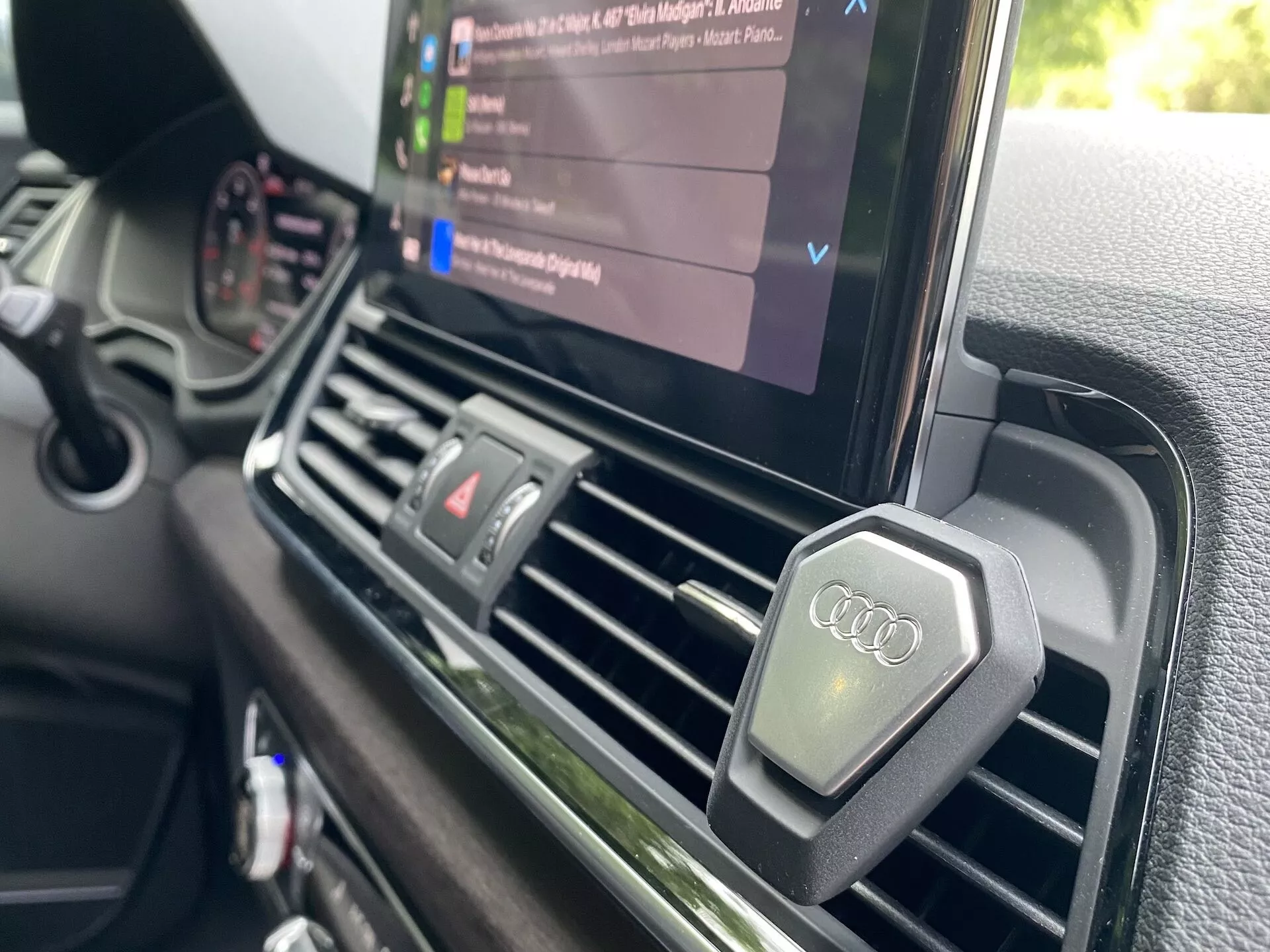 Zbliżenie z perspektywy kierowcy, pokazujące centralny kokpit 2020 Audi Q5 Sportback. Uwaga skupiona jest na centralnych nawiewach i ekranie systemu multimedialnego wyświetlającego odtwarzanie multimediów z listą plików audio, podkreślając możliwości multimedialne pojazdu. Nawiewy są prostokątne, o metalicznym wykończeniu i zawierają pokrętła z logo Audi. Deska rozdzielcza posiada teksturowaną, wysokiej jakości powierzchnię, zgodną z luksusowym pozycjonowaniem marki Audi, a brak widocznych ręcznych kontrolerów klimatyzacji sugeruje obecność automatycznego systemu klimatyzacji.
