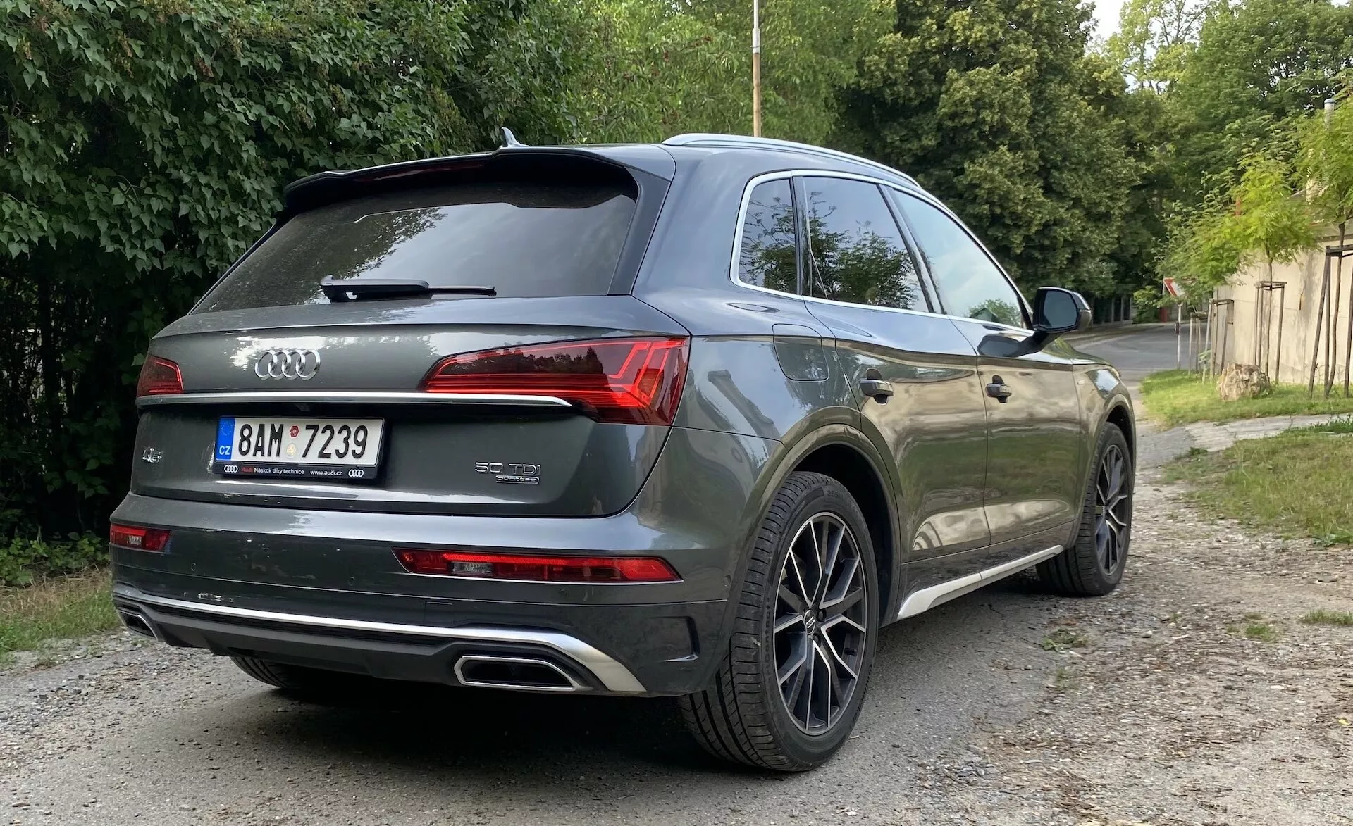 Widok pod kątem tylnym szarego SUV-a Audi Q5 Sportback 2020 zaparkowanego na żwirowej powierzchni obok zielonej roślinności. Obraz koncentruje się na tylnym i bocznym profilu samochodu, pokazując nowoczesne tylne światła LED, podwójne rury wydechowe, logo Audi pierścienie, oznaczenia modelu 'Q7' i wariantu '45 TDI', wieloszprychowe felgi aluminiowe, prywatne szyby, spojler dachowy i możliwe czujniki parkowania lub kamerę. Naturalne oświetlenie z miękkimi cieniami sugeruje wschód lub zachód słońca.