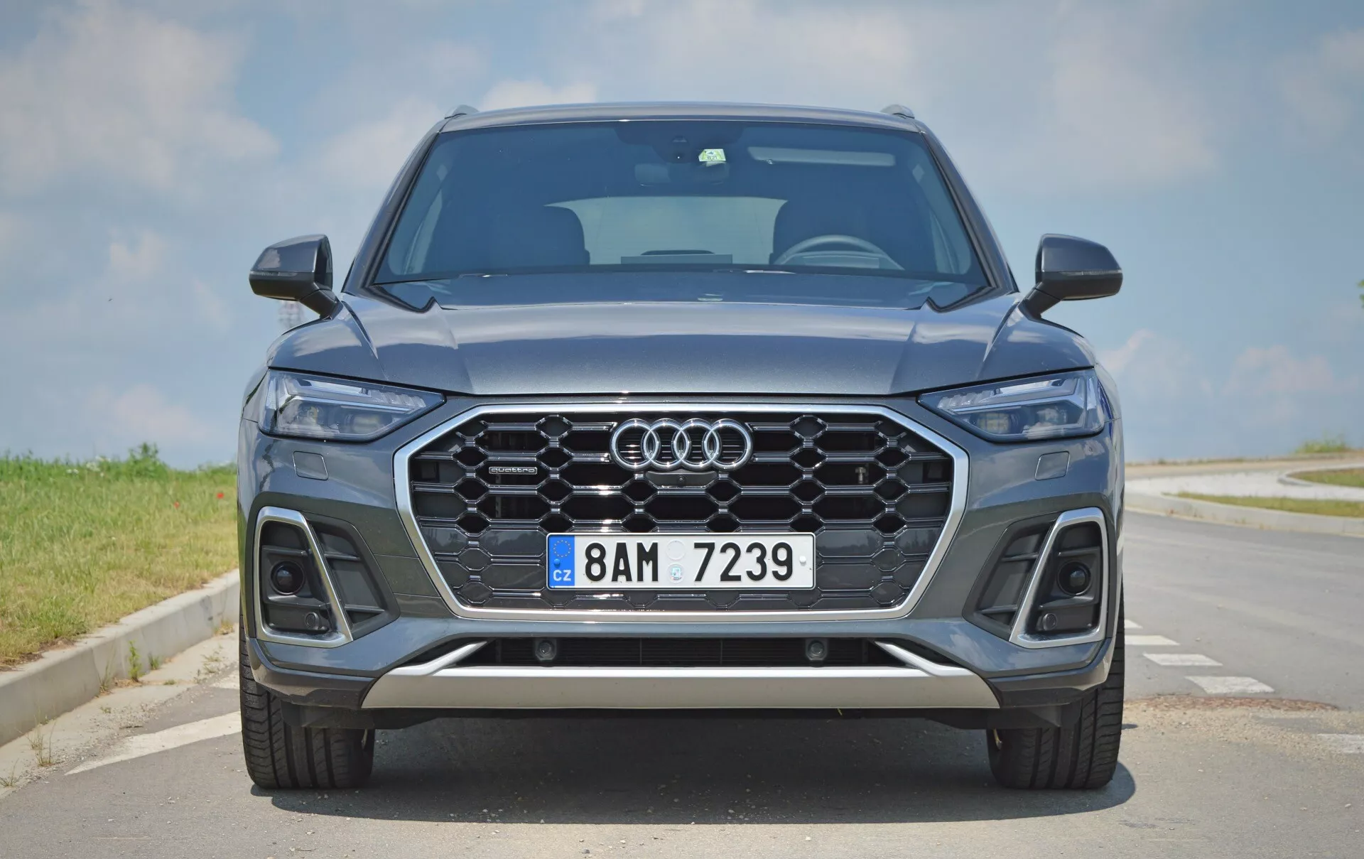 Widok z przodu nowoczesnego Audi Q5 2020 Sportback SUV prezentujący jego elegancki i symetryczny przód, z charakterystycznym grillem Audi, reflektorami LED, czujnikami parkowania, metalicznym niebieskim lakierem, felgami aluminiowymi i srebrnymi detalami wykończeniowymi. Scena ustawiona jest na brukowanej drodze pod czystym niebem.