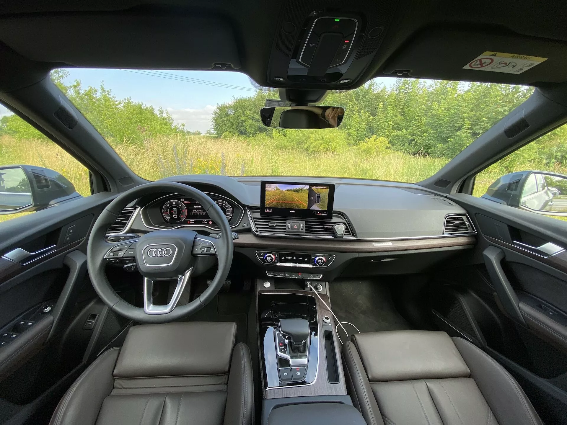Wnętrze Audi Q5 Sportback 2020 sfotografowane z perspektywy fotela kierowcy, prezentujące luksusowy i technologicznie zaawansowany kokpit. Obraz podkreśla czarne skórzane fotele, konsolę środkową z skórzanym podłokietnikiem, automatyczną dźwignię zmiany biegów otoczoną przyciskami sterującymi, poziomy ekran dotykowy systemu infotainment z nawigacją GPS, dwustrefową kontrolę klimatu, cyfrowy wyświetlacz kierowcy za kierownicą z logo Audi i zamontowanymi kontrolkami. Design charakteryzuje minimalizm, akcenty z matowego metalu i pełną widoczność przez szybę oraz słupek samochodu, co sugeruje, że jest zaparkowany na zewnątrz.