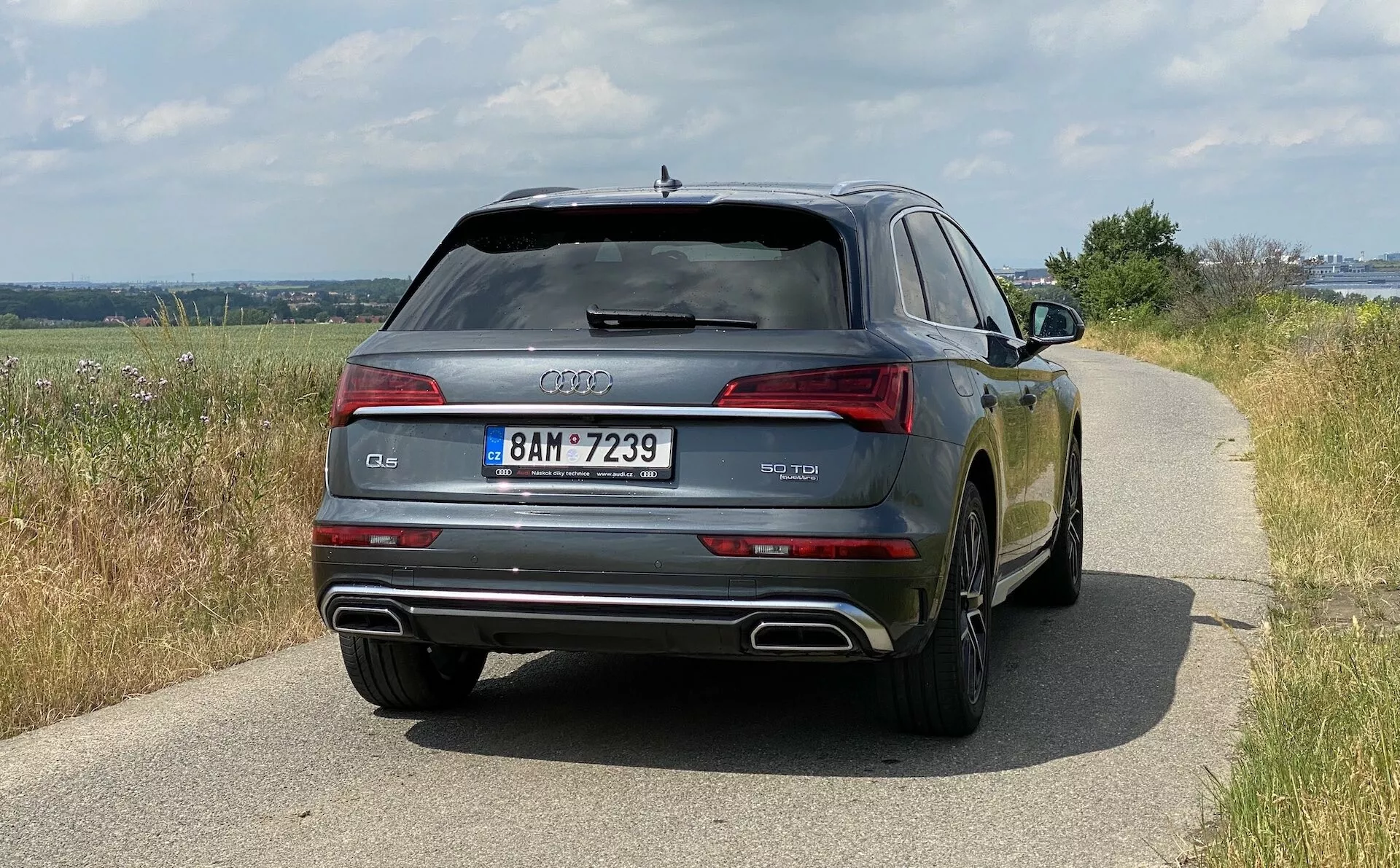Widok z tyłu Audi Q5 Sportback 2020, pokazujący szerokość pojazdu, w tym tylne światła, tylną szybę i podwójne rury wydechowe. Samochód, znajdujący się na drodze w świetle dziennym, ukazuje wiejskie otoczenie, a jego błyszczące wykończenie lakieru odbija otoczenie. Widoczne są szczegóły projektowe, takie jak pierścienie Audi, oznaczenie modelu i tablica rejestracyjna. Widoczne są również cechy technologiczne, takie jak światła LED, czujniki parkowania i tylna szyba odmrażająca.