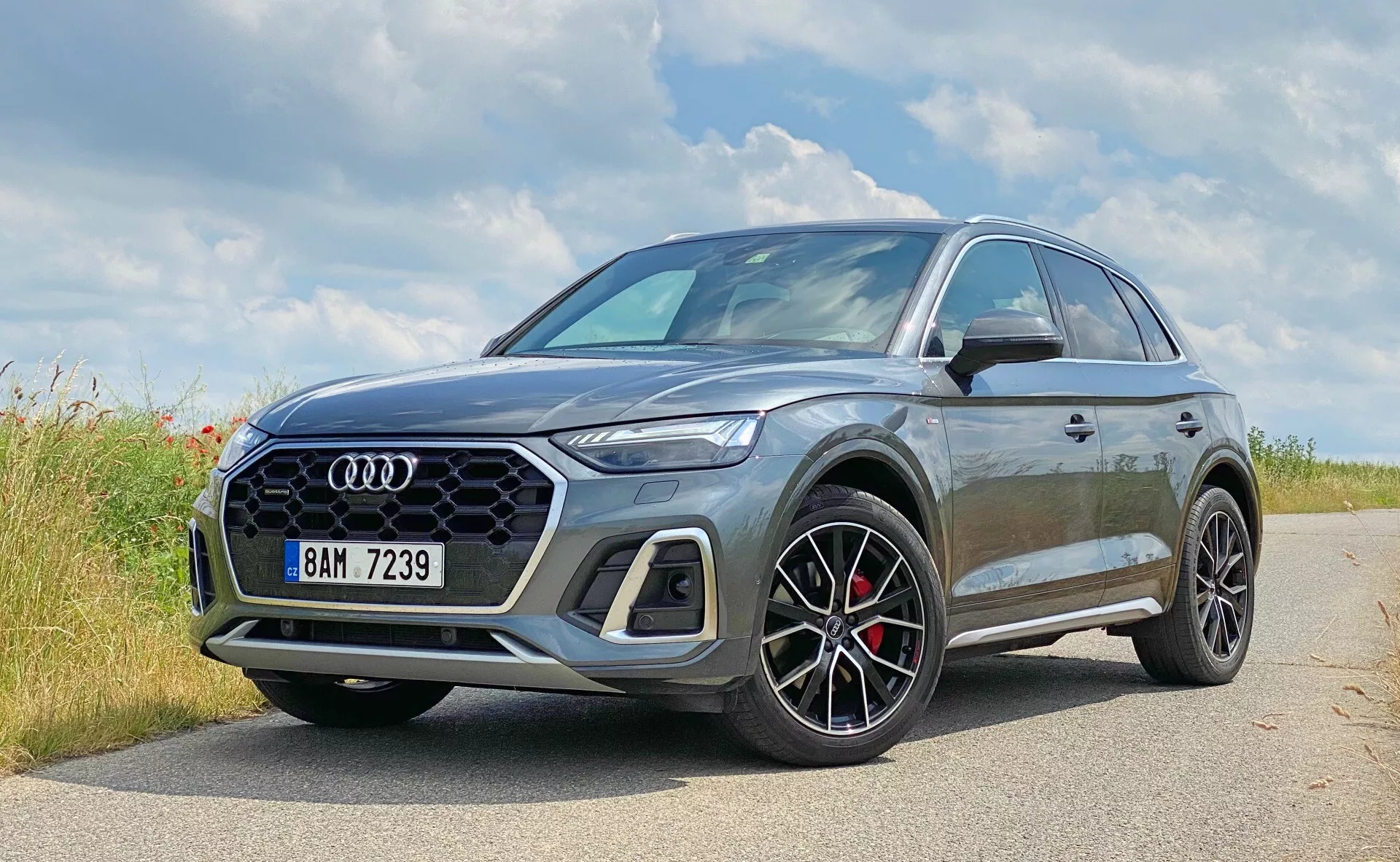 Obraz przedstawia nowoczesne SUV Audi Q5 Sportback 2020 zaparkowane na brukowanej bocznej drodze w naturalnym otoczeniu, prawdopodobnie wiejskim. Na pierwszym planie widoczne jest charakterystyczne logo Audi z czterema pierścieniami. Ten zbliżony, trzy-czwarte przedni widok podkreśla lewą przednią stronę i częściowy profil pojazdu, eksponując elementy designu, takie jak sześciokątna kratka wlotu powietrza z wzorem siatki, ostre kątowe reflektory LED oraz duże wieloramienne koła z czarnymi detalami i czerwonymi zaciskami hamulcowymi. Metaliczna, szara farba odbija światło, podkreślając kontury pojazdu. Nieco podniesione zawieszenie SUV-a sugeruje wszechstronność i lekkie zdolności terenowe, a lusterka boczne mają kontrastujący kolor, co może wskazywać na sportowy pakiet. Obraz oddaje luksusowy i sportowy design samochodowy z potencjalnie zaawansowanymi technologiami, których nie widać.