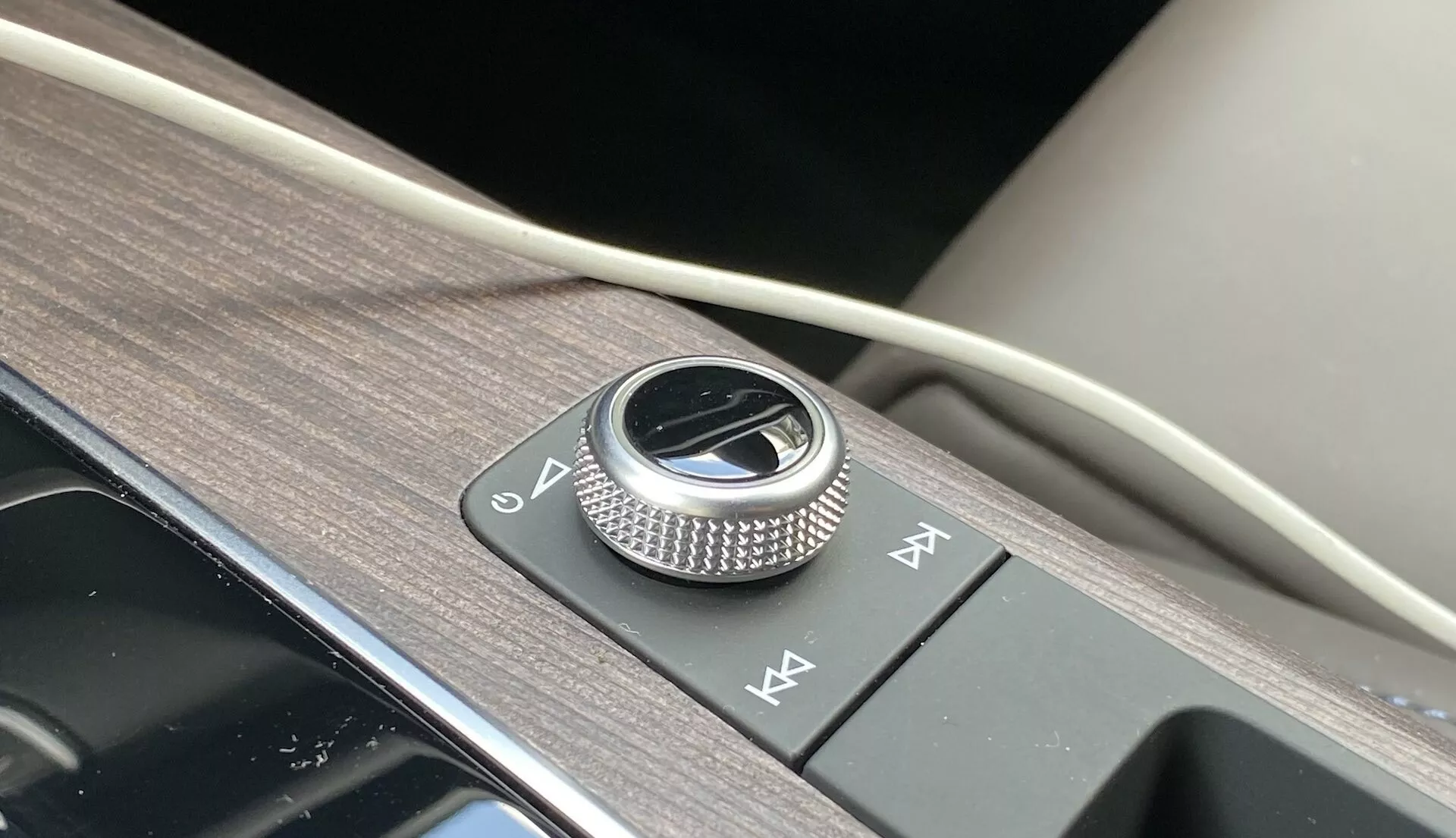 Zbliżenie konsoli środkowej Audi Q5 2020 Sportback, ukazujące metalowe pokrętło sterowania infotainment otoczone szczotkowanym metalem na lśniącym czarnym panelu. Obok znajdują się dwa przyciski szybkiego dostępu, wszystko na powierzchni z fakturą drewna z dodatkiem miękkiego materiału, co przykłada się do luksusowego designu wnętrza i zaawansowanych funkcji systemu infotainment.