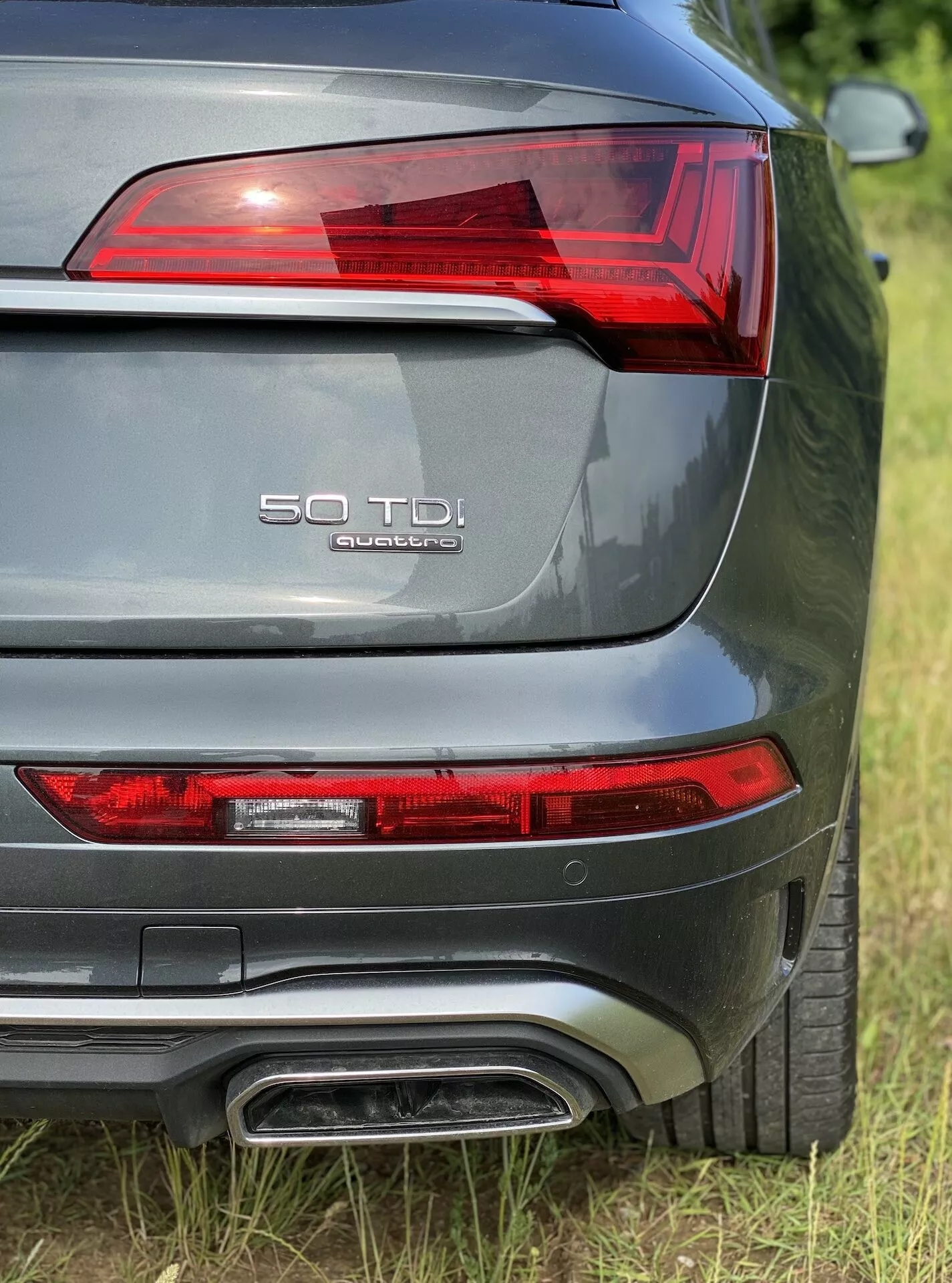 Zbliżenie na tylną sekcję Audi Q5 2020 Sportback z perspektywy trzech czwartych tylnej, pokazujące lewą tylną lampę z elementami LED, oznaczenie modelu '50 TDI' i 'quattro' na klapie bagażnika, podwójny układ wydechowy, metaliczny szary lakier i widoczny tylny czujnik parkowania.