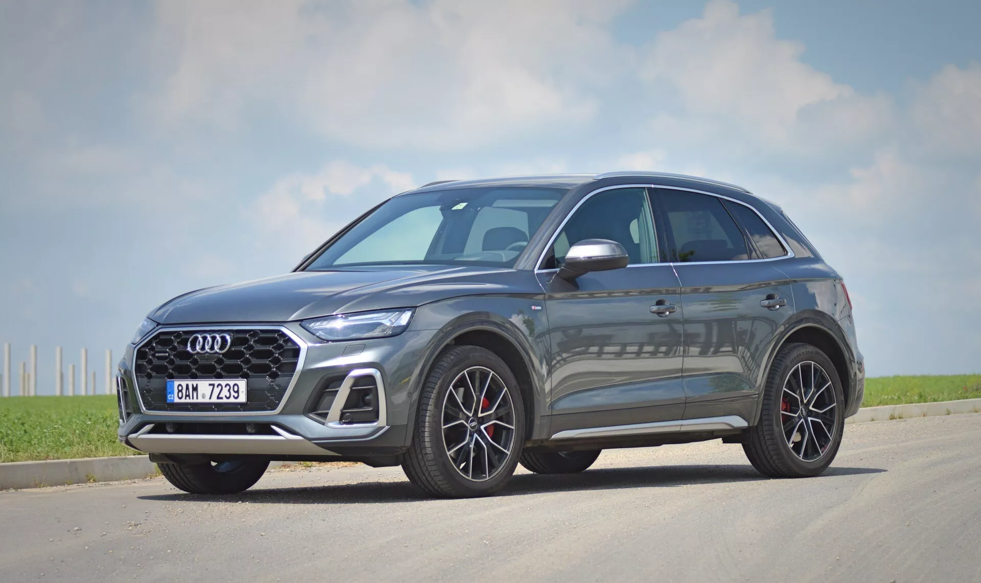 Obraz nowoczesnego crossover/SUV, modelu Audi Q5 Sportback 2020, stacjonującego na odcinku asfaltu. Samochód jest przedstawiony w widoku trzy-czwartych z przodu, ukazując zarówno przedni, jak i boczny profil. Wyraźny grill z lśniącym czarnym wzorem krzyżowym posiada centralnie umieszczony emblemat producenta. Smukłe, ostre reflektory LED i wieloramienne felgi aluminiowe zwiększają prestiżowy wygląd. Metaliczna farba odbija światło słoneczne, wskazując na wykończenie premium. Profil boczny ukazuje aerodynamiczne linie nadwozia i zewnętrzne lusterka boczne z zintegrowanymi kierunkowskazami. Okna mają chromowaną obwódkę kontrastującą z ciemnymi szybami, co może sugerować szkło przyciemniane. Samochód jest nieruchomy, umieszczony lekko na prawo od osi centralnej aparatu, a cienie sugerują, że jest rano lub po południu, pod czystym niebem z kilkoma chmurami w wiejskim lub półwiejskim otoczeniu.