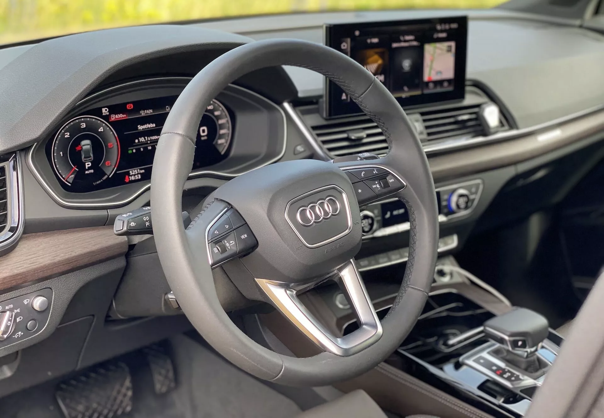 Luksusowe wnętrze Audi Q5 2020 Sportback z perspektywy kierowcy, ukazujące nowoczesną trójramienną kierownicę multifunkcyjną z zintegrowanymi kontrolkami, w pełni cyfrowy zestaw wskaźników oraz centralnie umieszczony ekran multimedialny wyświetlający nawigację. Wystrój wnętrza zawiera drewniane elementy, podkreślając poczucie luksusu i nowoczesności.