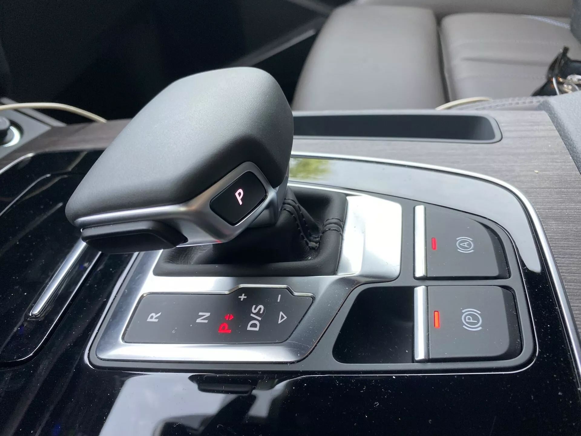 Zbliżenie na automatyczny selektor biegów w Audi Q5 2020 Sportback, umieszczony na konsoli środkowej, z projektem shift-by-wire w pozycji 'P' (Parkowanie), z opcjami 'R' (Wsteczny), 'N' (Neutralny) i 'D/S' (tryb jazdy/sportowy), plus znaki '+' i '-' dla trybu manualnego, przyciskiem 'Auto Hold' oraz elektronicznym hamulcem postojowym. Selektor jest otoczony wykończeniem w czarnym fortepianowym kolorze i chromowanymi akcentami, co wskazuje na luksusowe i technologicznie zaawansowane wnętrze.