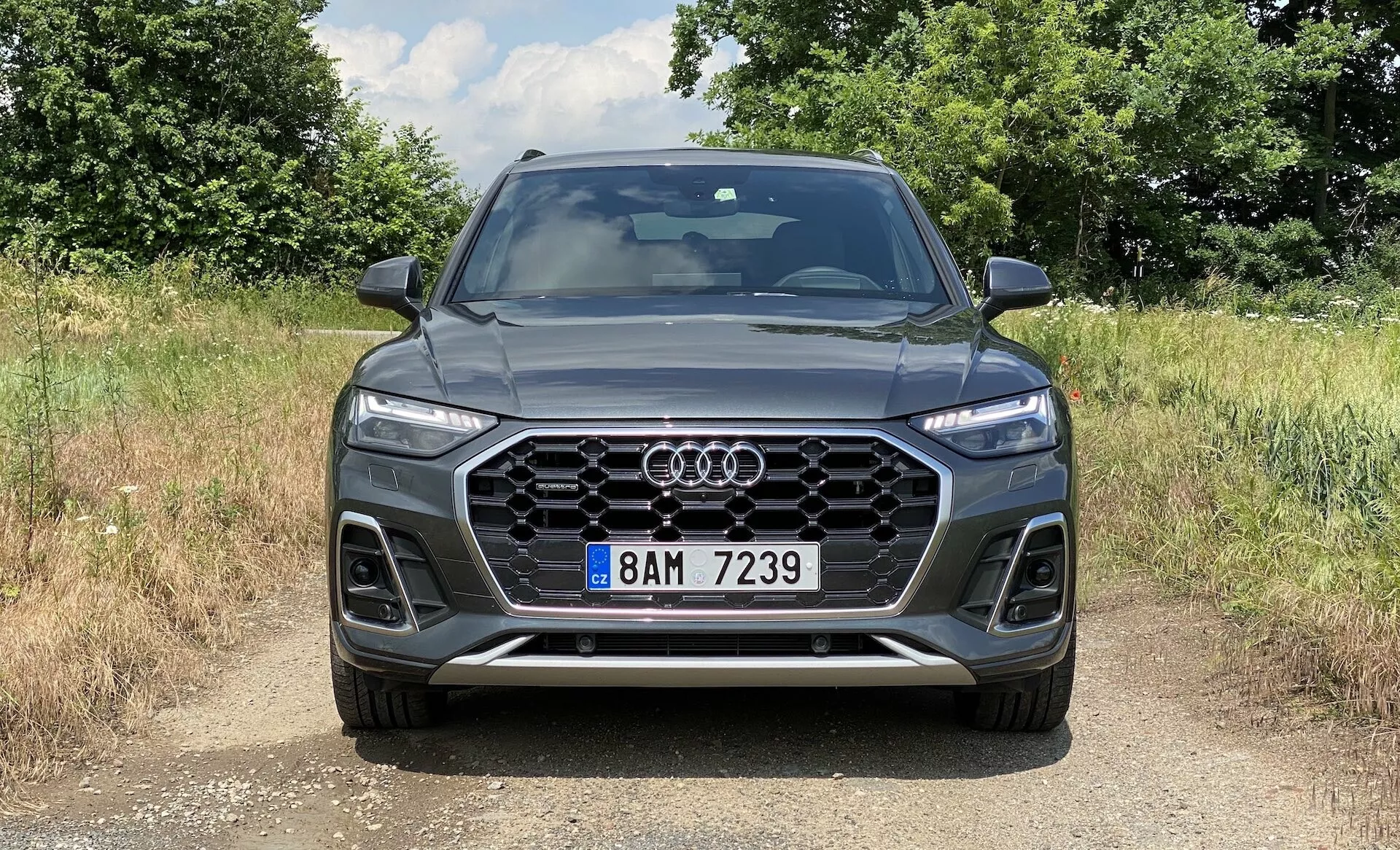 Widok z przodu na SUV Audi Q5 Sportback 2020 prezentujący wyraźną sześciokątną kratkę Singleframe z pionowymi chromowanymi listwami, centralnie umieszczonym znakiem Audi, ostre reflektory LED Matrix z charakterystycznymi światłami do jazdy dziennej, widoczne wloty powietrza z wzorzystym designem i przedni zderzak z wbudowanymi światłami przeciwmgielnymi i konturowanymi liniami. Samochód stoi na powierzchni z brudu lub żwiru, w naturalnym, wiejskim otoczeniu z promieniowaniem słonecznym rzucającym cienie, podkreślającym elegancki, sportowy wygląd i zaawansowane funkcje technologiczne.