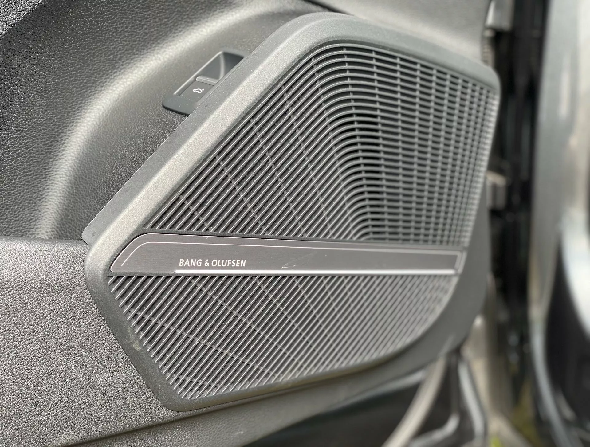 Zbliżenie głośnika premium BANG & OLUFSEN w panelu drzwiowym Audi Q5 Sportback 2020. Delikatny wzór siatki kratki jest wyraźnie widoczny, podczas gdy tło i pierwszy plan są rozmyte. Nad kratką znajduje się mała trójkątna sekcja, widoczna z boku i lekko przedniego kąta, prawdopodobnie zawierająca tweeter. Obraz podkreśla luksusową jakość systemu audio pojazdu.