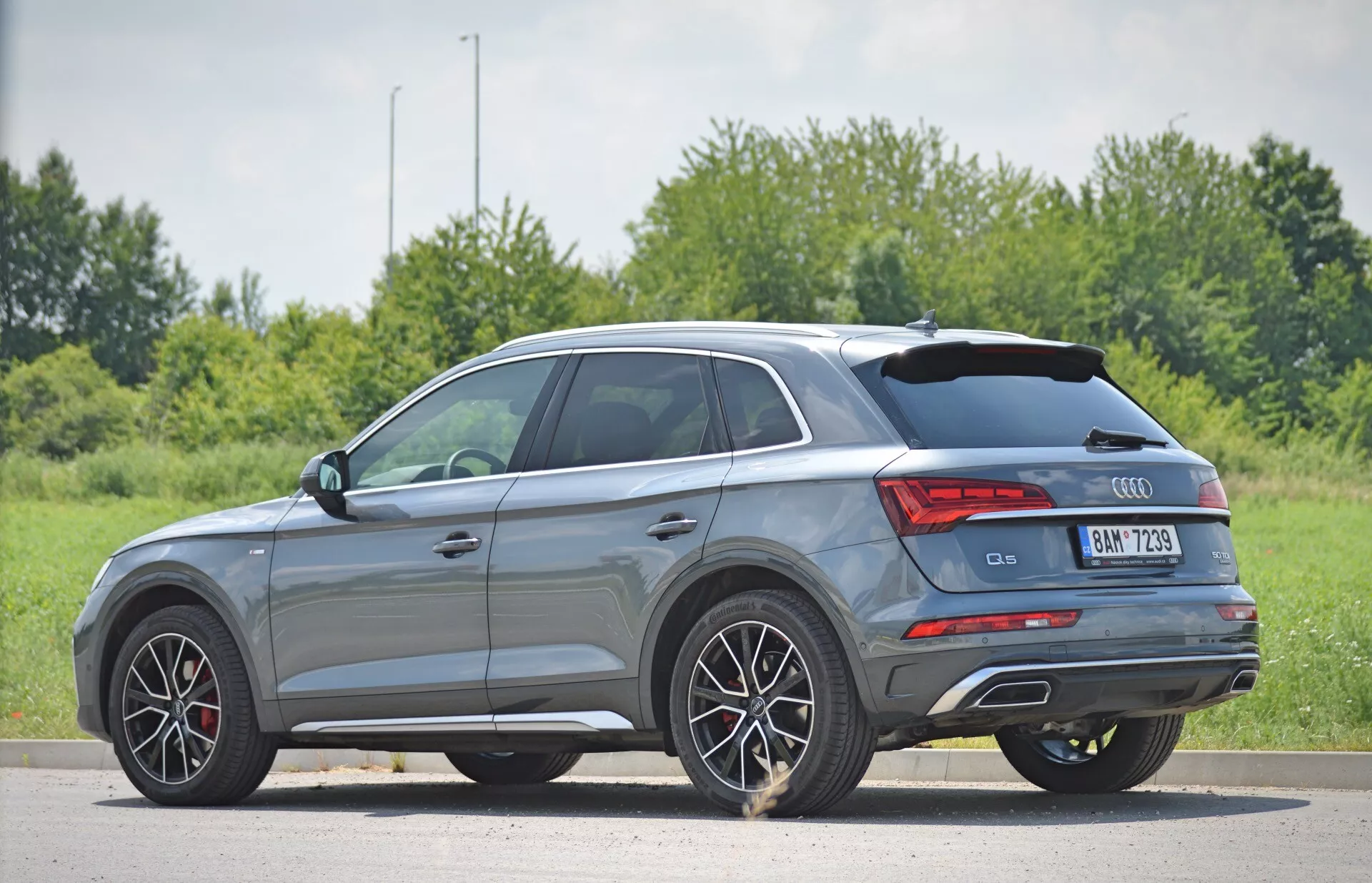 Szary SUV Audi Q5 Sportback z roku modelowego 2020 jest pokazany w trzech czwartych z tyłu, podkreślając eleganckie linie nadwozia, wieloramienne felgi aluminiowe, tylne światła LED, podwójne końcówki wydechu i opadającą tylną linię dachu. Samochód zaparkowany jest na równej powierzchni z naturalnym tłem trawy i drzew, a naturalne światło podkreśla sportowe elementy designu i kompaktowy wygląd średniej wielkości SUV-a.