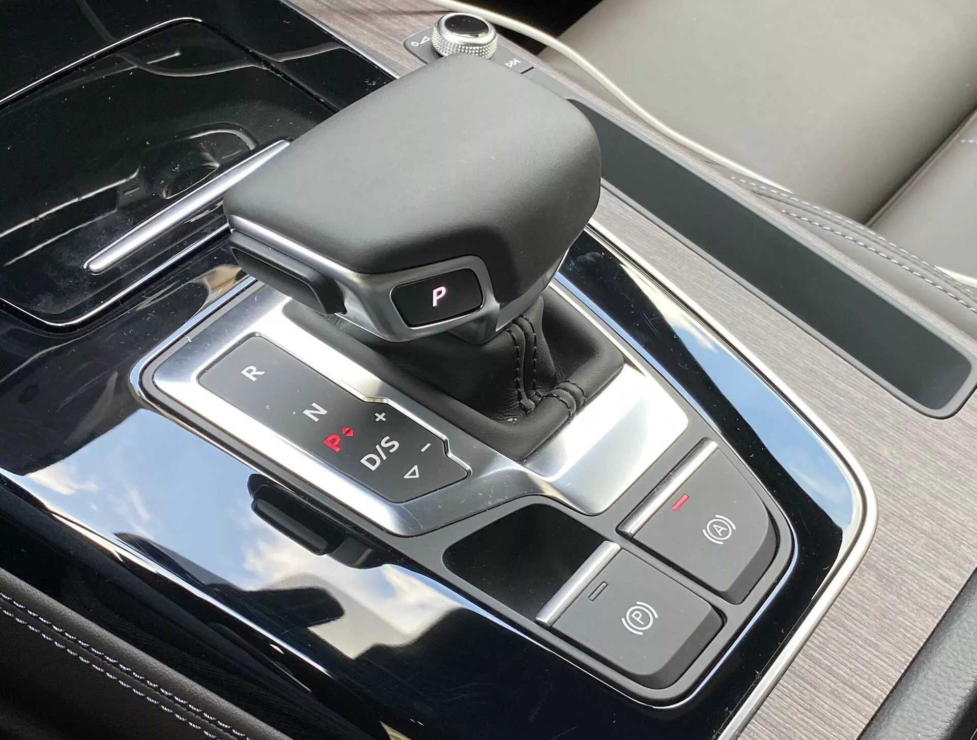 Widok z góry na konsolę centralną w Audi Q5 Sportback 2020, podkreślający elegancki obszar zmiany biegów z automatycznym selektorem skórzanym, podświetlanymi wskaźnikami biegów oraz sąsiadującymi przyciskami do automatycznego hamulca postojowego i funkcji Auto Start/Stop. Konsola posiada wykończenie przypominające czarny fortepian z srebrnymi akcentami, odzwierciedlając luksusowy wystrój wnętrza i nowoczesną technologię.