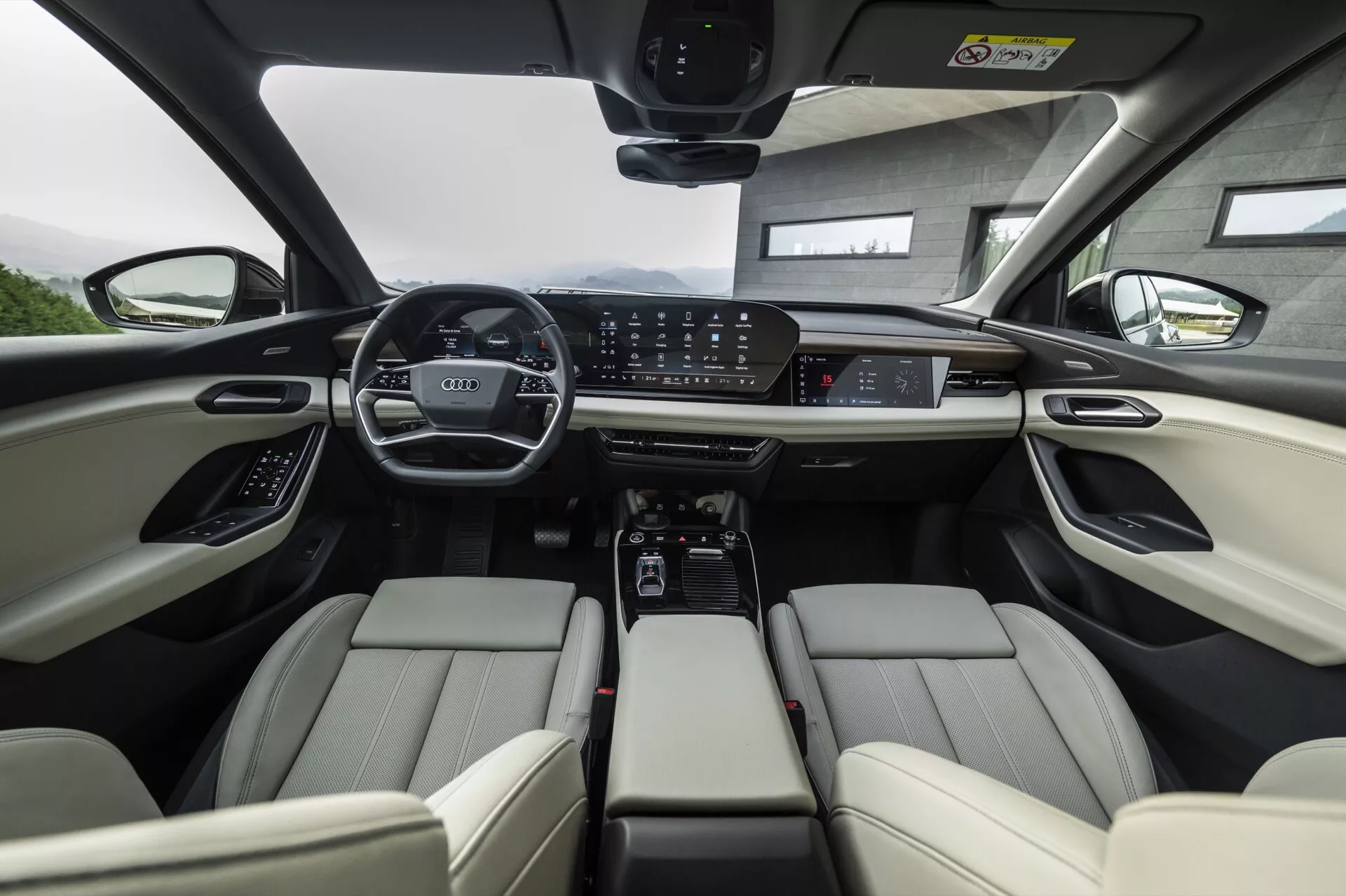 Wnętrze Audi Q6 e-tron 2024 z perspektywy tylnego siedzenia, skupiając się na przednim rzędzie. Jasne, skórzane przednie fotele wyposażone są w regulowane zagłówki i elektroniczne sterowanie. Deska rozdzielcza prezentuje szeroki cyfrowy panel wskaźników oraz duży centralny ekran dotykowy do systemu info-rozrywki, otoczony przyciskami sterującymi klimatyzacją i trybami jazdy. Konsola środkowa zawiera uchwyty na kubki, miejsce do przechowywania oraz selektor automatycznej skrzyni biegów. Kierownica jest wyposażona w liczne elementy sterujące, a panoramiczne okno dachowe sugeruje obfite światło naturalne. Ogólnie obraz podkreśla zaawansowane technologie i luksusowe cechy samochodu.