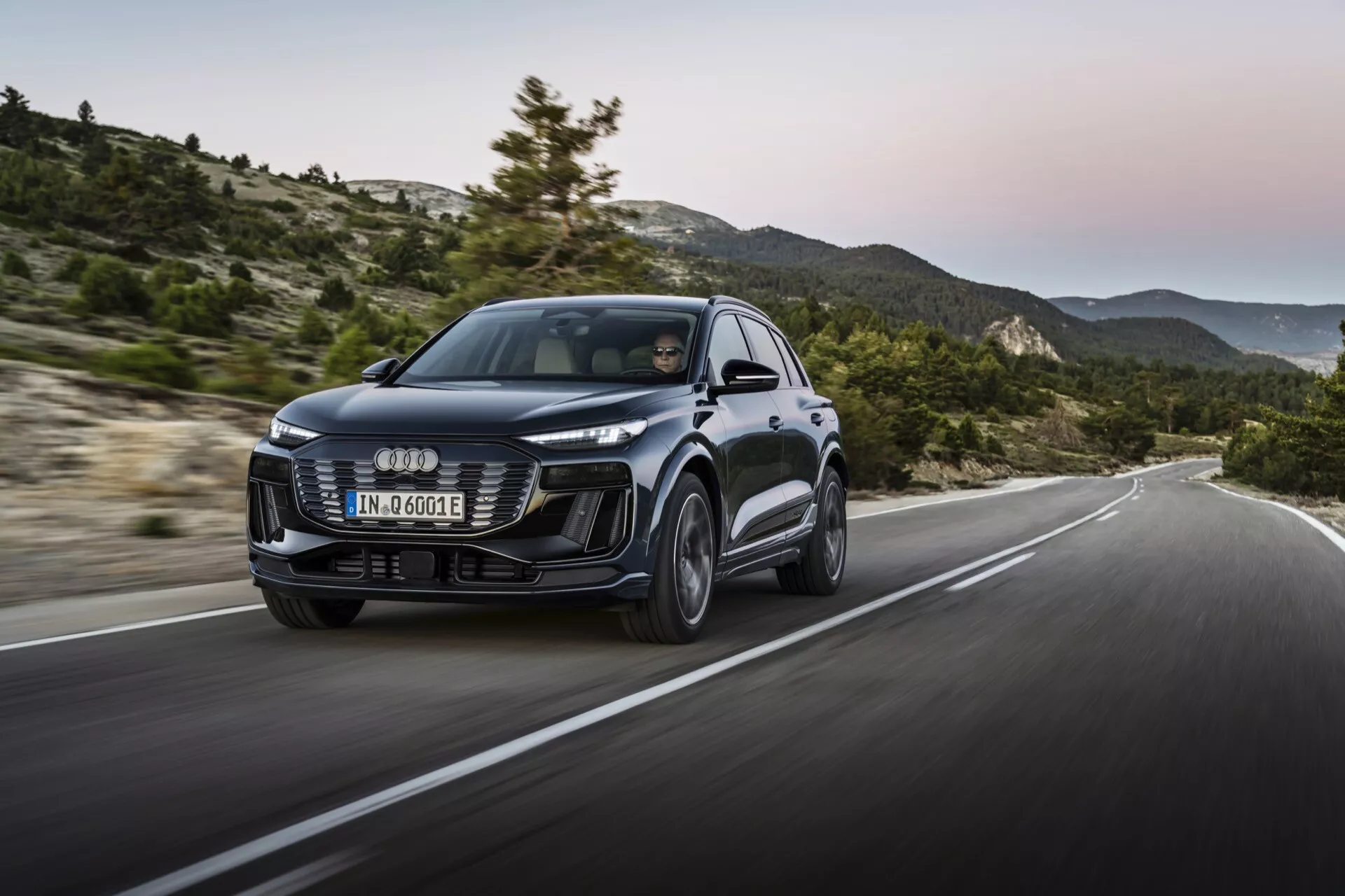 Audi Q6 e-tron 2024 z nowoczesnym designem, dużą przednią kratką, eleganckimi reflektorami LED i rzeźbionymi liniami nadwozia. Widok z trzech czwartych od przodu z dynamicznym ujęciem w ruchu na pagórkowej drodze, podkreślającym wydajność i prowadzenie.