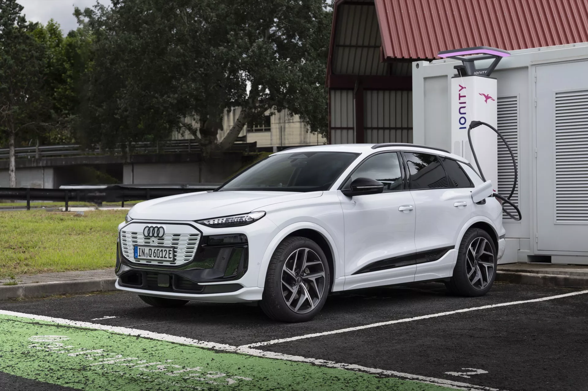 Widok z przodu i z boku na biały elektryczny SUV Audi Q6 e-tron 2024, podkreślający przedni grill, reflektory LED, linie aerodynamiczne, wieloramienne koła i boczny profil. Pojazd zaparkowany jest na wyznaczonym miejscu do ładowania pojazdów elektrycznych z widocznym portem ładowania i ładowarką IONITY w tle. Obraz kontrastuje design samochodu z ciemnym, pochmurnym niebem i mokrym chodnikiem, ukazując nowoczesną technologię pojazdów elektrycznych i zaawansowane oświetlenie.