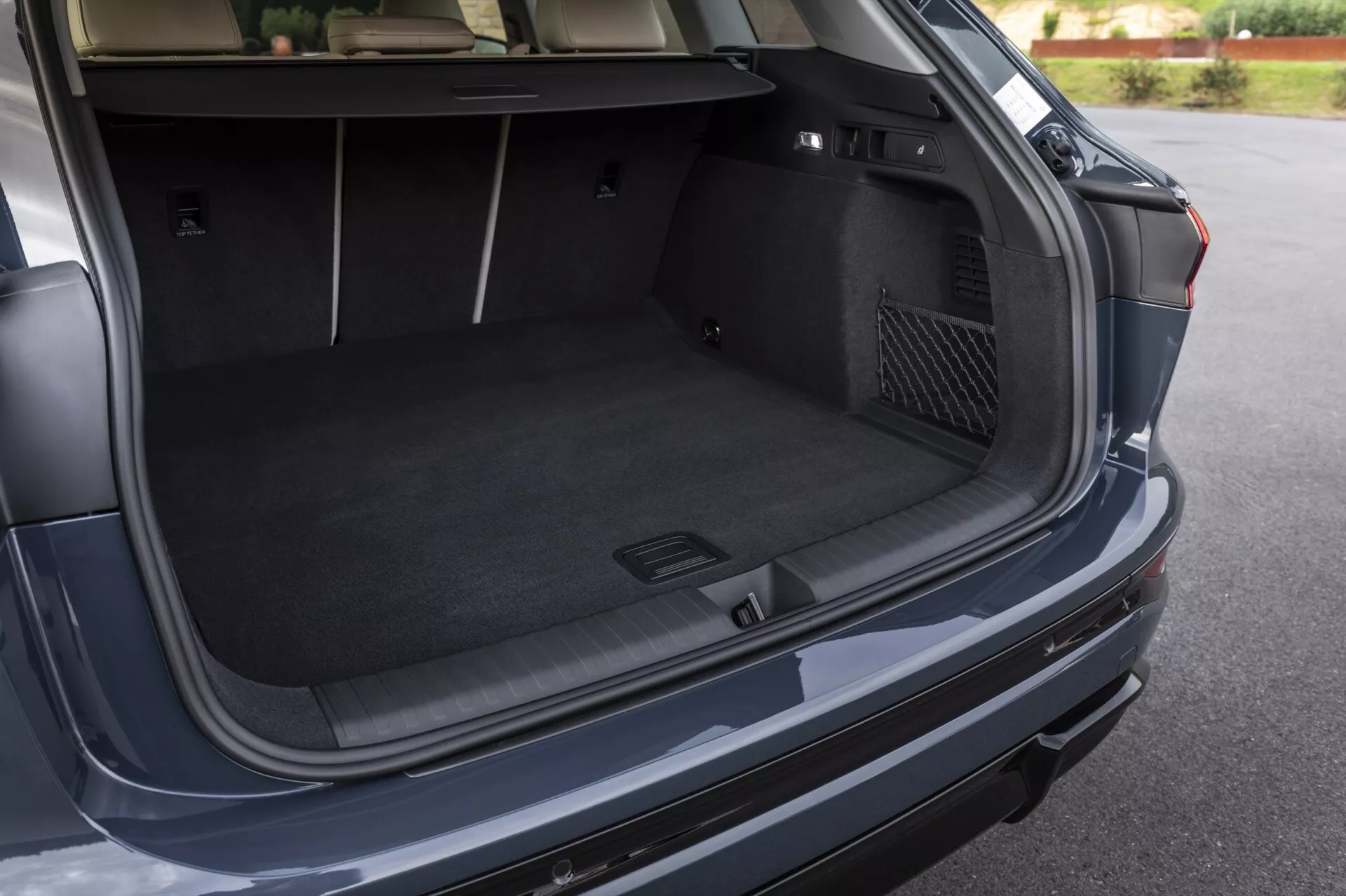 Obraz przedstawia otwarty bagażnik nowoczesnego pojazdu Audi Q6 e-tron 2024, uchwycony z kąta nieco powyżej i na prawo od tyłu samochodu. Ten punkt widzenia zapewnia wyraźny widok na przestronny, wykładany wykładziną obszar bagażnika, podkreślając jego głębokość i szerokość. Notatki godne uwagi obejmują formowany schowek po lewej stronie, przeznaczony na drobne przedmioty lub sprzęt awaryjny oraz kieszeń z siatki po prawej stronie. Na pokrywie bagażnika widoczny jest możliwy przycisk automatycznego zamykania. Tylni zderzak i częściowy widok prawej tylnej lampy sugerują design zewnętrzny pojazdu.