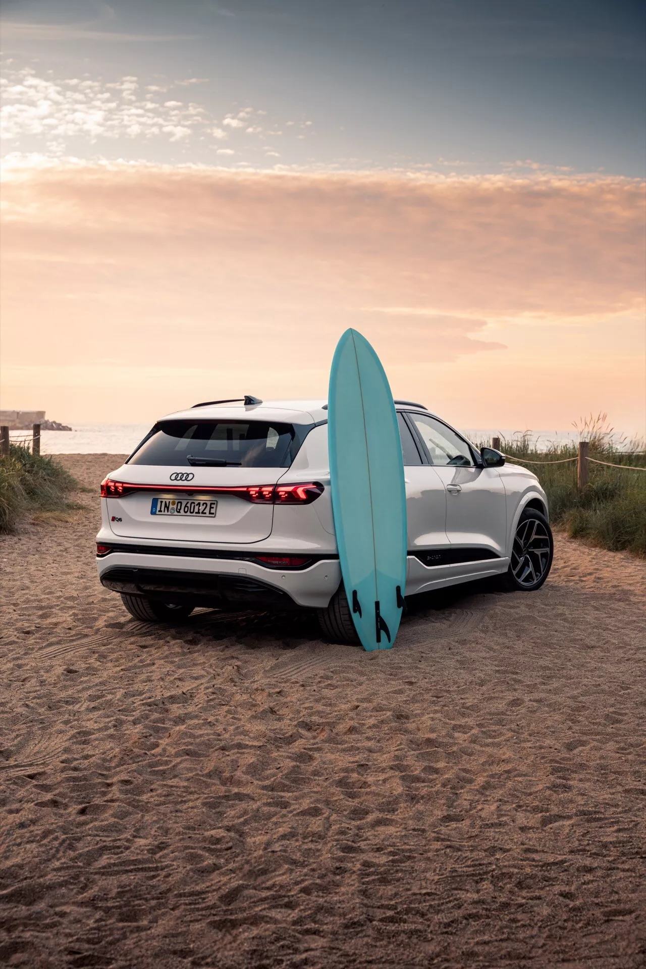 Obraz przedstawia białe Audi Q6 e-tron 2024 SUV zaparkowane na piaszczystym terenie plażowym z deską surfingową. Fotografowane pod kątem trzech czwartych od tyłu, uwidacznia eleganckie linie samochodu, tylne światła LED, czujniki parkowania, kamerę cofania oraz wieloramienne alufelgi. Odbicie zachodzącego słońca podkreśla zaawansowaną jakość lakieru, a deska surfingowa oparta o samochód akcentuje element przygodowego stylu życia.