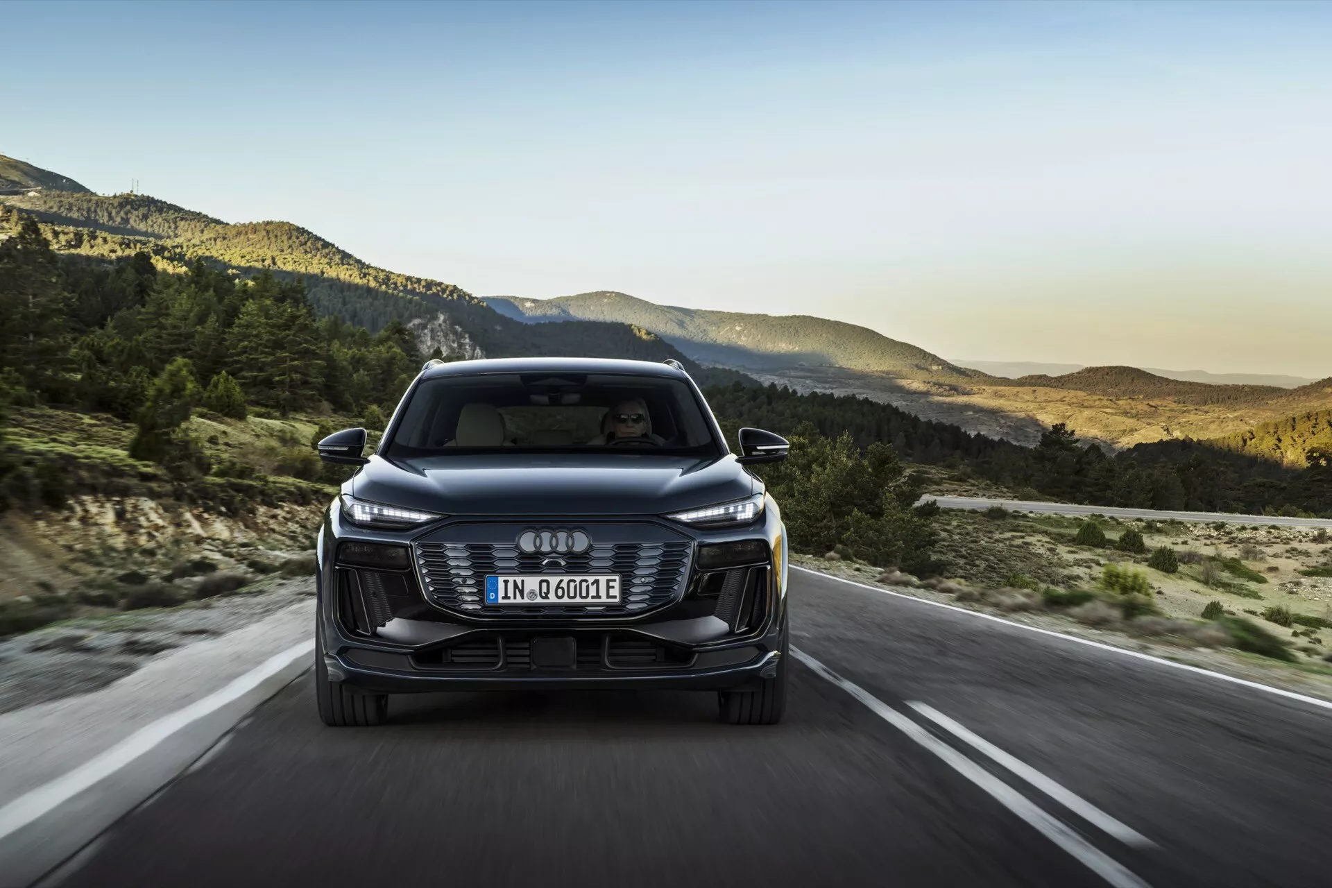 Widok czołowy Audi Q6 e-tron 2024, prezentujący przedni grill, reflektory i zderzak z logo czterech złączonych pierścieni. Europejska tablica rejestracyjna jest zamontowana na grillu, a oznaczenie 'e-tron' wskazuje na jego elektryczny/hybrydowy charakter. Samochód jedzie otwartą drogą z górskim krajobrazem w tle, sugerując ustawienie późnego popołudnia. Cechy obejmują eleganckie reflektory LED, estetyczny grill, czujniki/kamery wspomagania kierowcy, aerodynamiczne lusterka i efektywny design.