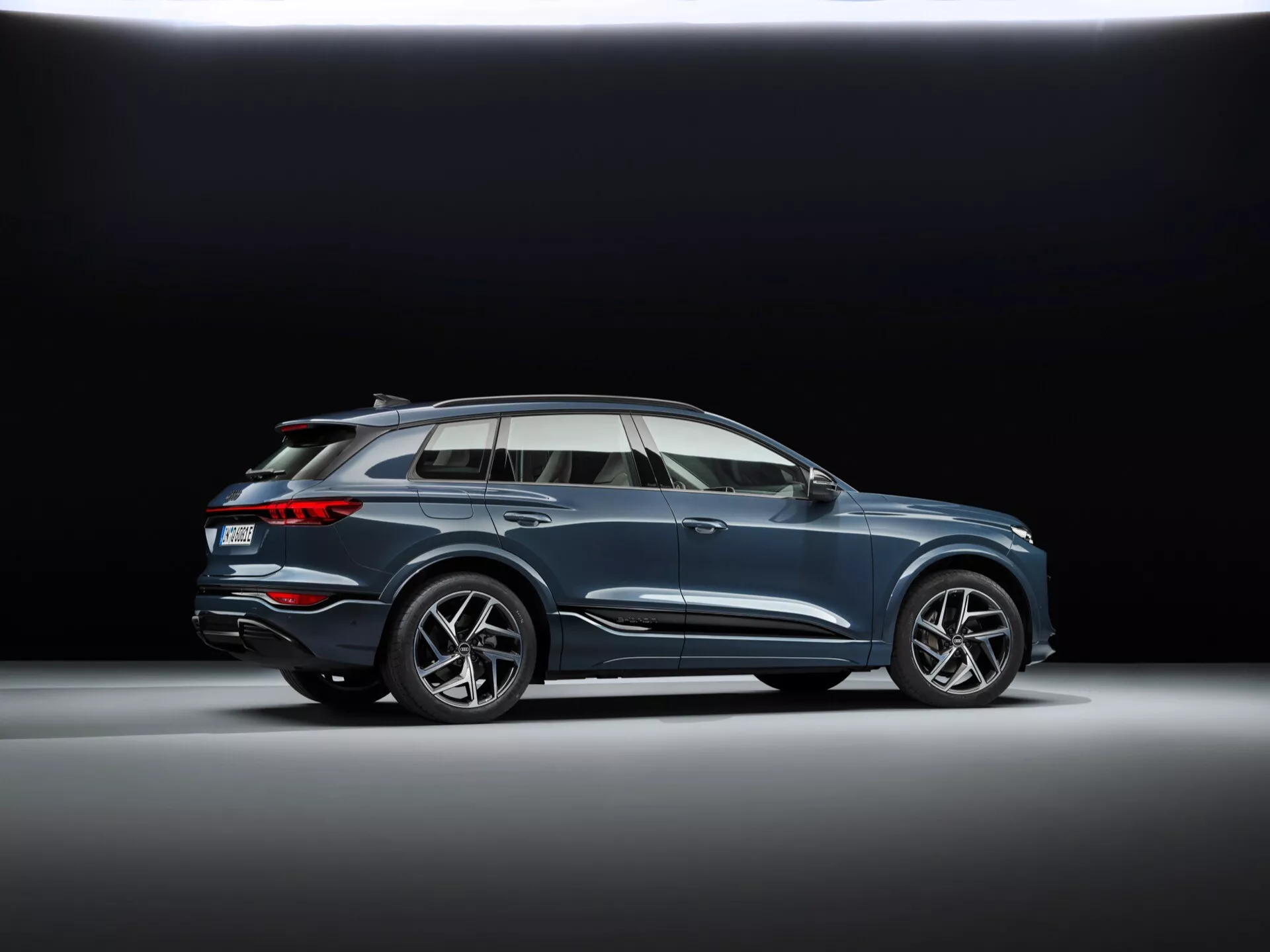 Niskokątny profilowy strzał Audi Q6 e-tron 2024 w studio z ciemnym tłem, z sportowymi wieloramiennymi felgami, płynną linią nadwozia z charakterystyczną linią, aerodynamicznymi lusterkami bocznymi z wbudowanymi kierunkowskazami, chromowanymi obwódkami okien, panoramicznym dachem, relingami dachowymi, umiarkowaną wysokością jazdy i podwójnym układem wydechowym flush.