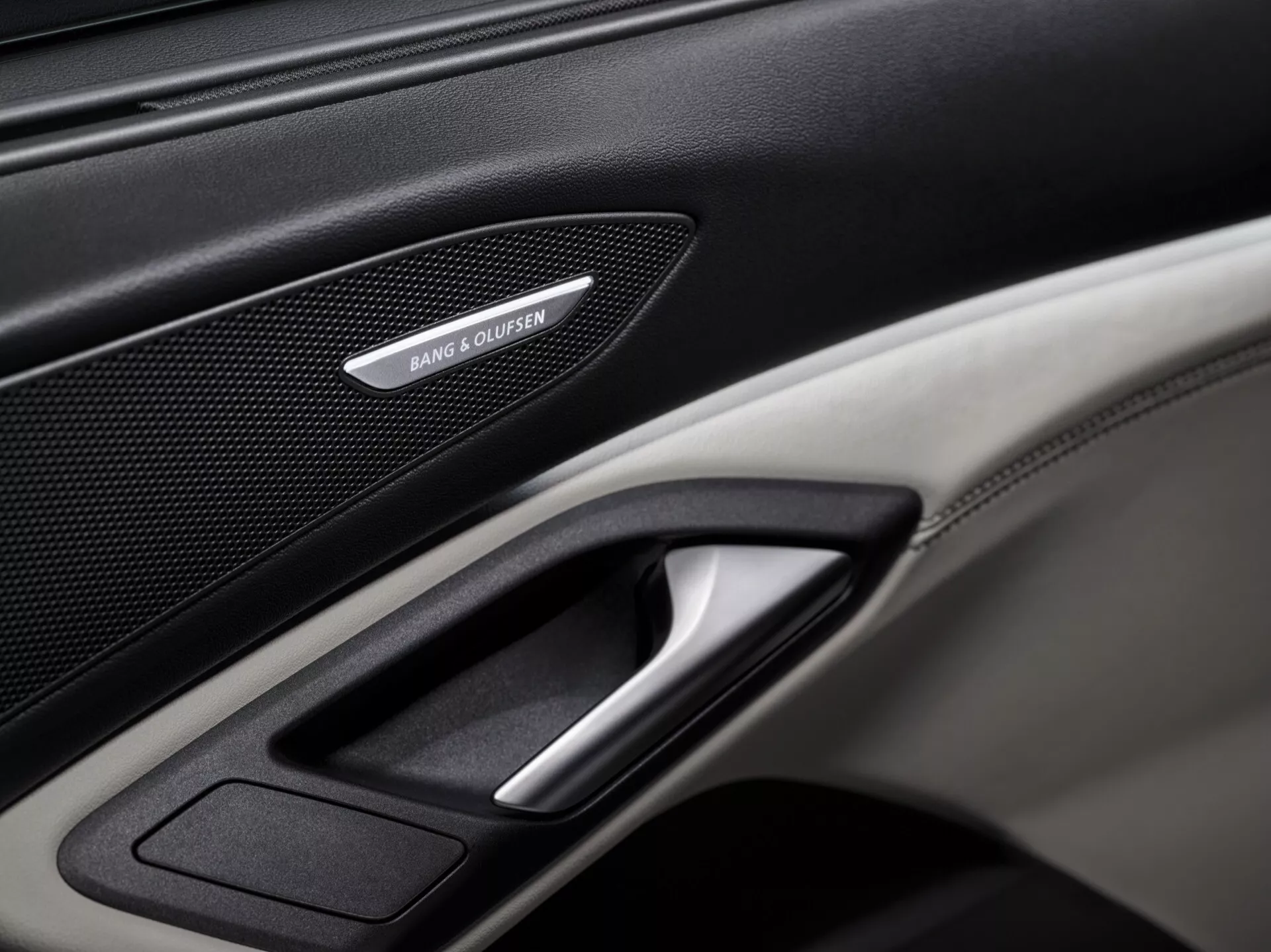 Zbliżenie wnętrza panelu drzwiowego Audi Q6 e-tron 2024 GF, podkreślające luksusowe funkcje takie jak kratka głośnika Bang & Olufsen, chromowana dźwignia drzwi oraz kombinacja skórzanych i karbonowych tekstur. Dobrze oświetlone zdjęcie skupia się na szczegółach panelu, ukazując luksusowe i technologiczne elementy pojazdu.
