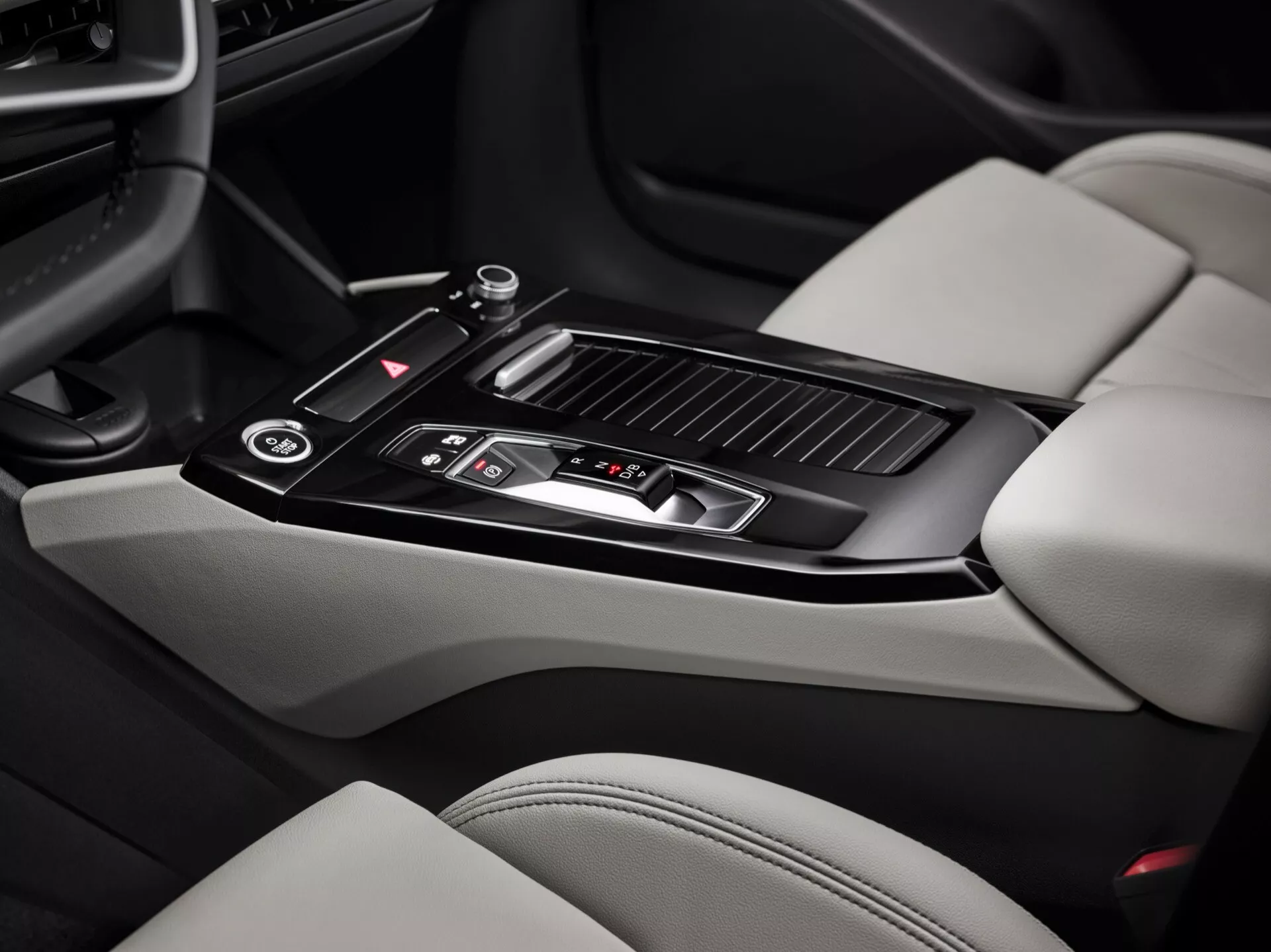 Zdjęcie wnętrza Audi Q6 e-Tron 2024, skupiające się na obszarze konsoli środkowej. Obraz, zrobiony z perspektywy pasażera, ukazuje błyszczącą czarną wajchę zmiany biegów z metalicznymi detalami, kilka przycisków kontrolnych, małe miejsce na przechowywanie z suwaną pokrywą, dwa uchwyty na kubki, tapicerowany skórą podłokietnik, kratki wentylacyjne klimatyzacji i części jasnokolorowych skórzanych siedzeń z kontrastującymi ciemniejszymi sekcjami. Funkcje konsoli środkowej ukazują nowoczesny design i luksusowe elementy.