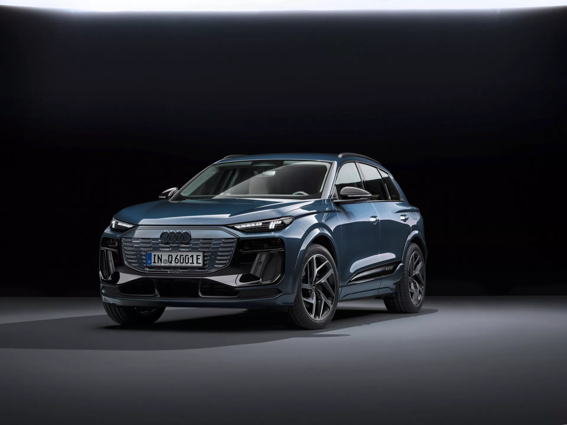 Obraz przedstawia nowoczesne SUV Audi Q6-e-tron 2024 z widoku z trzech czwartych przodu, podkreślając zarówno profil przedni, jak i boczny. Kluczowymi identyfikatorami są cztery pierścienie znaku Audi na grillu oraz odznaka 'e-tron', wskazująca na model elektryczny. Ujęcie wykonano z niskiej perspektywy, podkreślając sylwetkę pojazdu i prezentując linie projektowe, muskularne nadkola i cechy aerodynamiczne. Samochód jest umieszczony w centrum kadru, stoi na gładkiej refleksyjnej powierzchni z miękkim, rozproszonym oświetleniem i ciemnym gradientowym tłem, co akcentuje design pojazdu. Widoczne cechy technologiczne to reflektory LED, światła dzienne (DRL), wyświetlacz cyfrowy i skomplikowanie zaprojektowane felgi aluminiowe. Ogólna dopracowana prezentacja sugeruje fotografię promocyjną, która podkreśla technologię, design i luksusowe aspekty pojazdu.