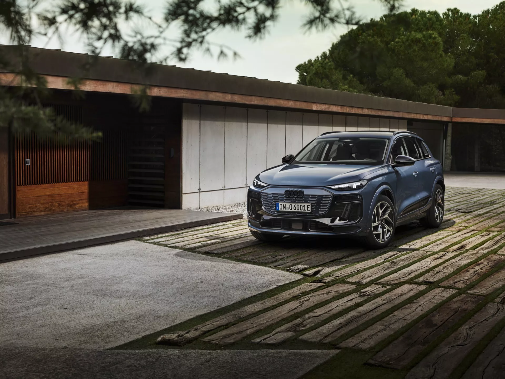 Widok z przodu za trzy czwarte Audi Q6 E-Tron 2024 SUV z eleganckimi reflektorami LED, wyraźnym grillem i dużym emblematem zaparkowanym na wzorzystej nawierzchni. Luksusowy budynek i drzewa w tle podkreślają luksus i ekskluzywność.