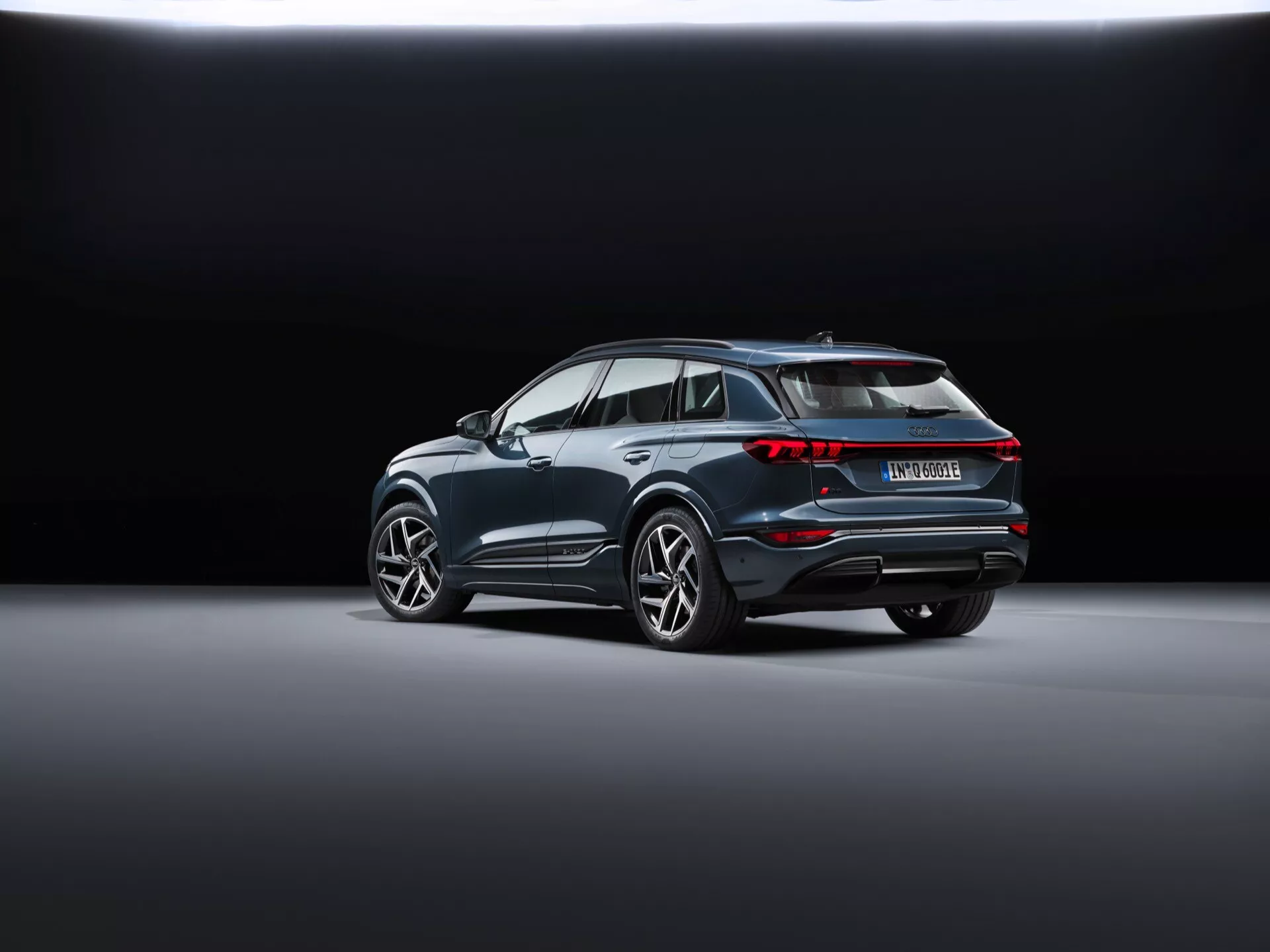 Widok tylno-trzyczwarty Audi Q6 e-tron 2024 (gf) przedstawia nowoczesne SUV ustawione po przekątnej na gładkiej, odblaskowej podłodze showroomu. Tło przechodzi z ciemnego na jasny, podkreślając pojazd. Widoczne cechy to połączone tylne światła, charakterystyczny spojler dachowy, wyraźne nadkola mieszczące duże felgi aluminiowe, chromowana linia wokół okien, tylni zderzak z odblaskami i czujnikami oraz podwójny układ wydechowy sugerujący osiągi, co może wskazywać na model hybrydowy lub elektryczny.