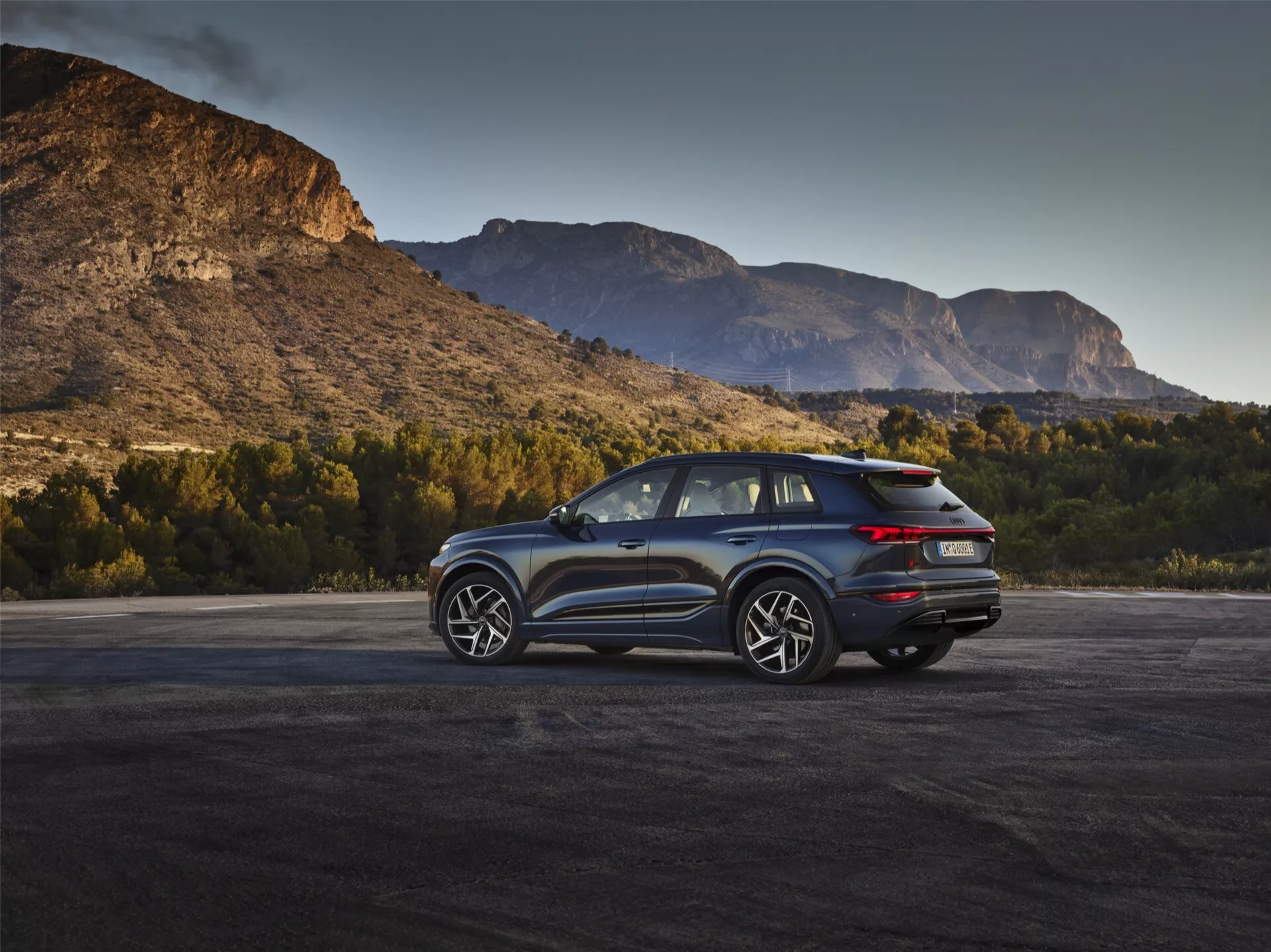 Obraz przedstawia nowoczesne SUV Audi Q6 e-Tron 2024 w plenerze z malowniczym górskim tłem podczas świtu lub zmierzchu. Kąt tylno-boczny podkreśla charakterystyczne tylne światła LED, czysty design tylnej klapy z możliwą kamerą cofania oraz podwójny układ wydechowy. Profil boczny ukazuje wieloramienne felgi aluminiowe, duże okna i skośną linię dachu, co sugeruje sportową lub luksusową wersję z możliwymi cechami aerodynamicznymi. Lusterka boczne mogą mieć zintegrowaną technologię monitorowania martwego pola. Naturalne światło podkreśla atrakcyjność pojazdu, wskazując na jego przywiązanie do przygód i zdolności.