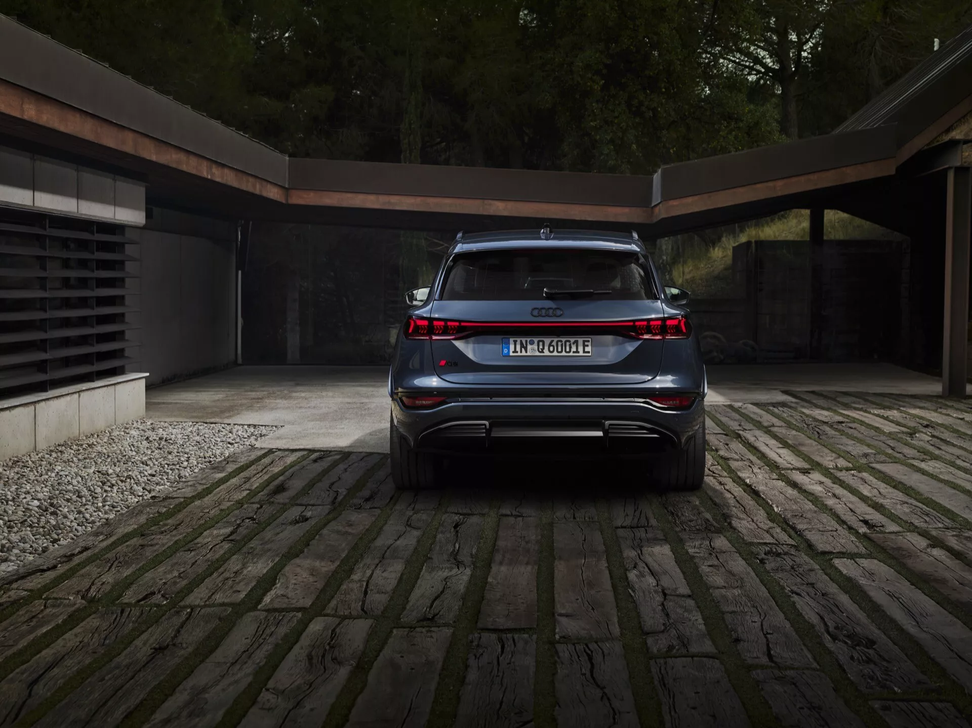 Widok z tyłu Audi Q6 e-tron 2024 podkreślający jego czysty, elegancki i nowoczesny design. Widoczne kluczowe cechy to ciągły pas świetlny łączący oba tylne światła, oznaczenia modelu i elektrycznego układu napędowego, zintegrowany tylny zderzak ze sportowym dyfuzorem, nachylona do przodu tylna szyba sugerująca SUV-a lub crossovera, ukryte końcówki wydechu i umieszczona centralnie tablica rejestracyjna. Samochód jest zaparkowany na brukowanym podjeździe z oznaką współczesnego garażu w tle, co wskazuje na luksusowy i ekologiczny styl życia.
