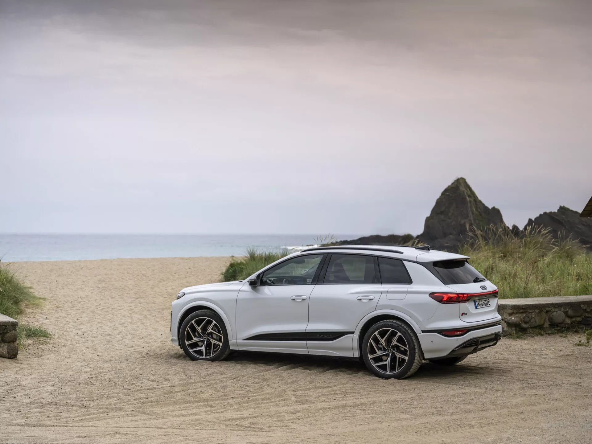 Biały SUV Audi Q6 e-tron 2024 zaparkowany na drodze gruntowej z plażowym tłem, uchwycony z trzech czwartych tylnego widoku. Obraz podkreśla nowoczesne elementy designu pojazdu, w tym tylne światła, tylny zderzak, stylowe koła i smukłe linie. Większy prześwit i ogólna postawa wskazują na przydatność do różnych terenów. Miękkie, równomierne oświetlenie sugeruje pochmurny dzień lub bliskość wschodu/zachodu słońca, podkreślając przygodowy nastrój. Widoczne są nowoczesne felgi, tylne spojler i tylne światła LED, sygnalizujące zaawansowaną technologię i sportowy charakter.