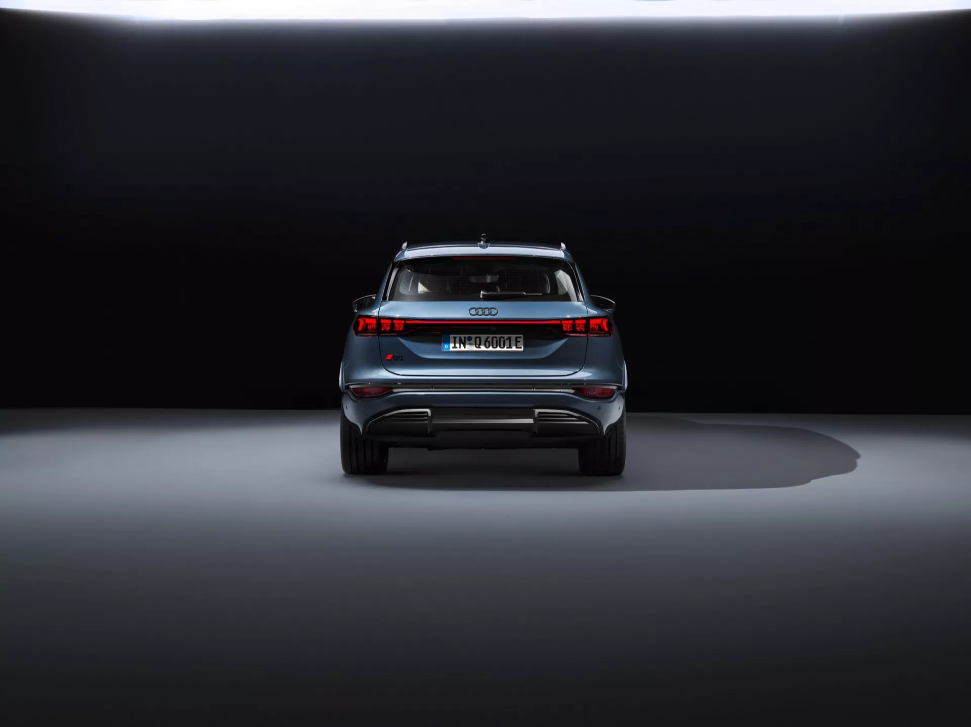Widok z tyłu Audi Q6 e-tron 2024 na wystawie z dramatycznym oświetleniem. Zdjęcie z poziomu ziemi podkreśla nowoczesne tylne światła LED, brak rur wydechowych, pełnowymiarowy pasek świetlny, europejską tablicę rejestracyjną, spojler dachowy i elementy odblaskowe, wskazując, że jest to luksusowy pojazd elektryczny lub hybrydowy