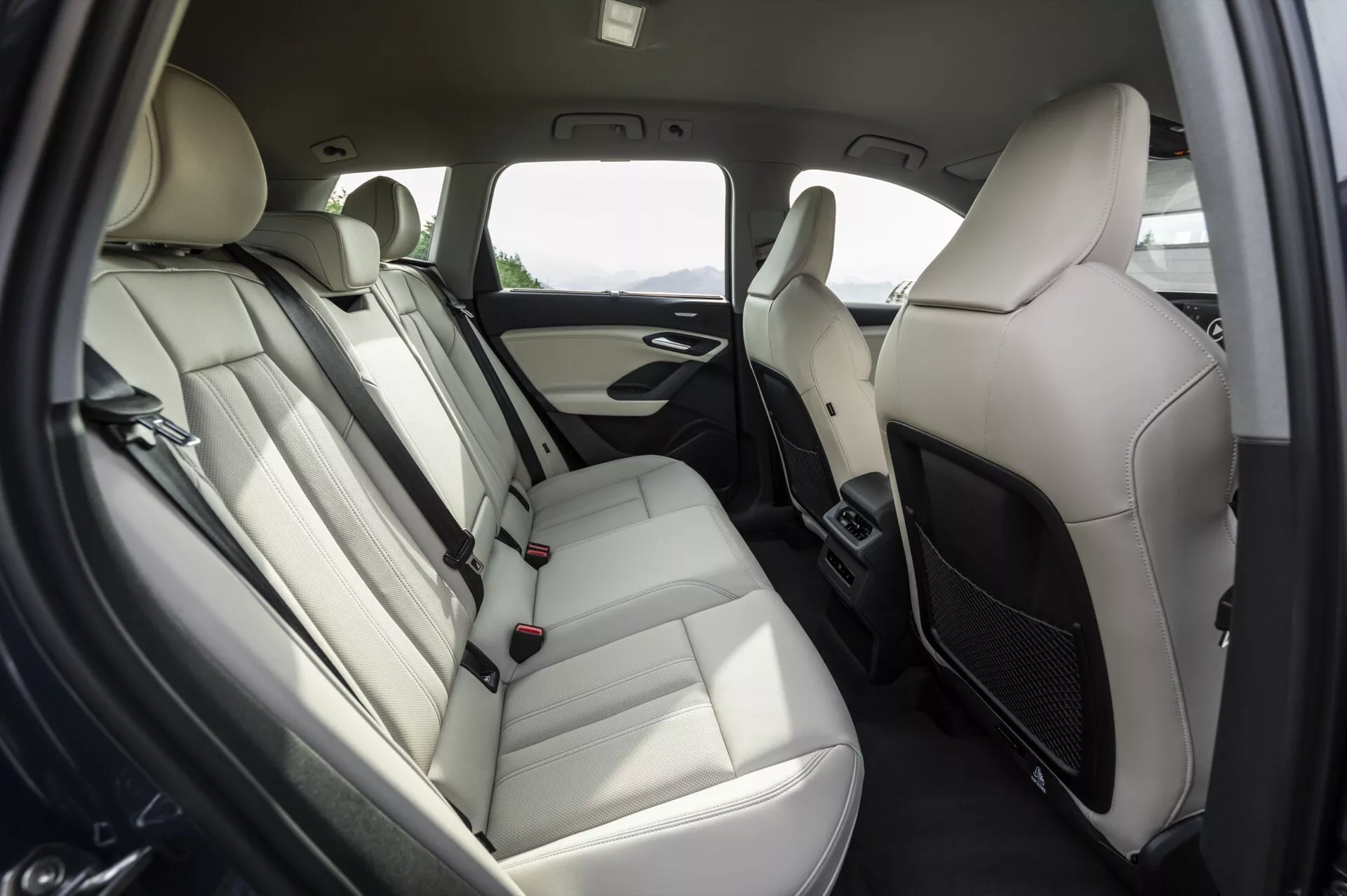 Wnętrze tylnego przedziału pasażerskiego Audi Q6 e-tron 2024 z naciskiem na układ tylnych siedzeń. Ławki pokryte jasną skórą z zagłówkami, podłokietnikiem i widocznymi pasami bezpieczeństwa z klamrami. Panel drzwi wyposażony w kratkę głośników, uchwyt i prawdopodobne sterowanie oknem. Dwukolorowy schemat kolorów z jasnymi i ciemnymi akcentami. Kratki klimatyzacji między przednimi siedzeniami wskazują na kontrolę klimatu dla pasażerów tylnego siedzenia. Nowoczesny, luksusowy, wygodny design dla pasażerów.