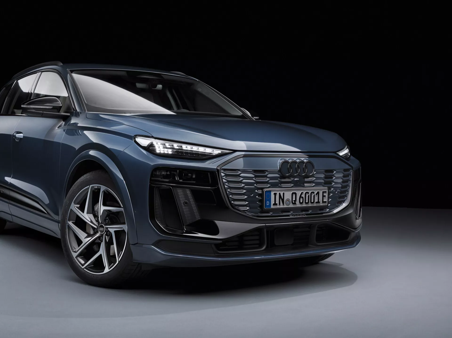 Obraz przedstawia nowoczesne Audi Q6 e-tron 2024 w scenerii studyjnej, ukazane z widoku w trzech czwartych z przodu, który daje wyraźne wrażenie projektowania pojazdu z przodu i z boku. Jasno widoczny jest lewy przedni narożnik samochodu, w tym przedni grill z wzorem plastra miodu i centralnie umieszczonym emblematem Audi, skomplikowane przednie światła LED, dodatkowe wloty powietrza lub czujniki pod reflektorami, sportowy zderzak przedni, duże koło z lekkich stopów z oponą o niskim profilu oraz kontur lusterka bocznego. Obraz podkreśla insignię modelu elektrycznego ('e') i uwydatnia jego cechy projektowe i technologiczne. Tło jest proste i ciemne, co zapewnia brak rozpraszania uwagi od samego pojazdu.