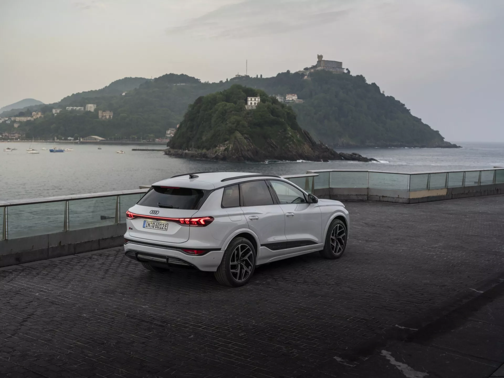 Trzy-czwartowy widok tylnej części nowoczesnego białego SUV-a Audi Q6 e-tron 2024 zaparkowanego na nadmorskiej esplanadzie o zmierzchu, prezentującego poziome tylne światła LED, znaczek producenta, tylnią wycieraczkę, pięcioramienne felgi aluminiowe, lusterka z wbudowanymi kierunkowskazami, relingi dachowe oraz rzeźbione linie nadwozia na tle spokojnego krajobrazu z małą wyspą i budynkiem, podkreślając połączenie użyteczności i wydajności.