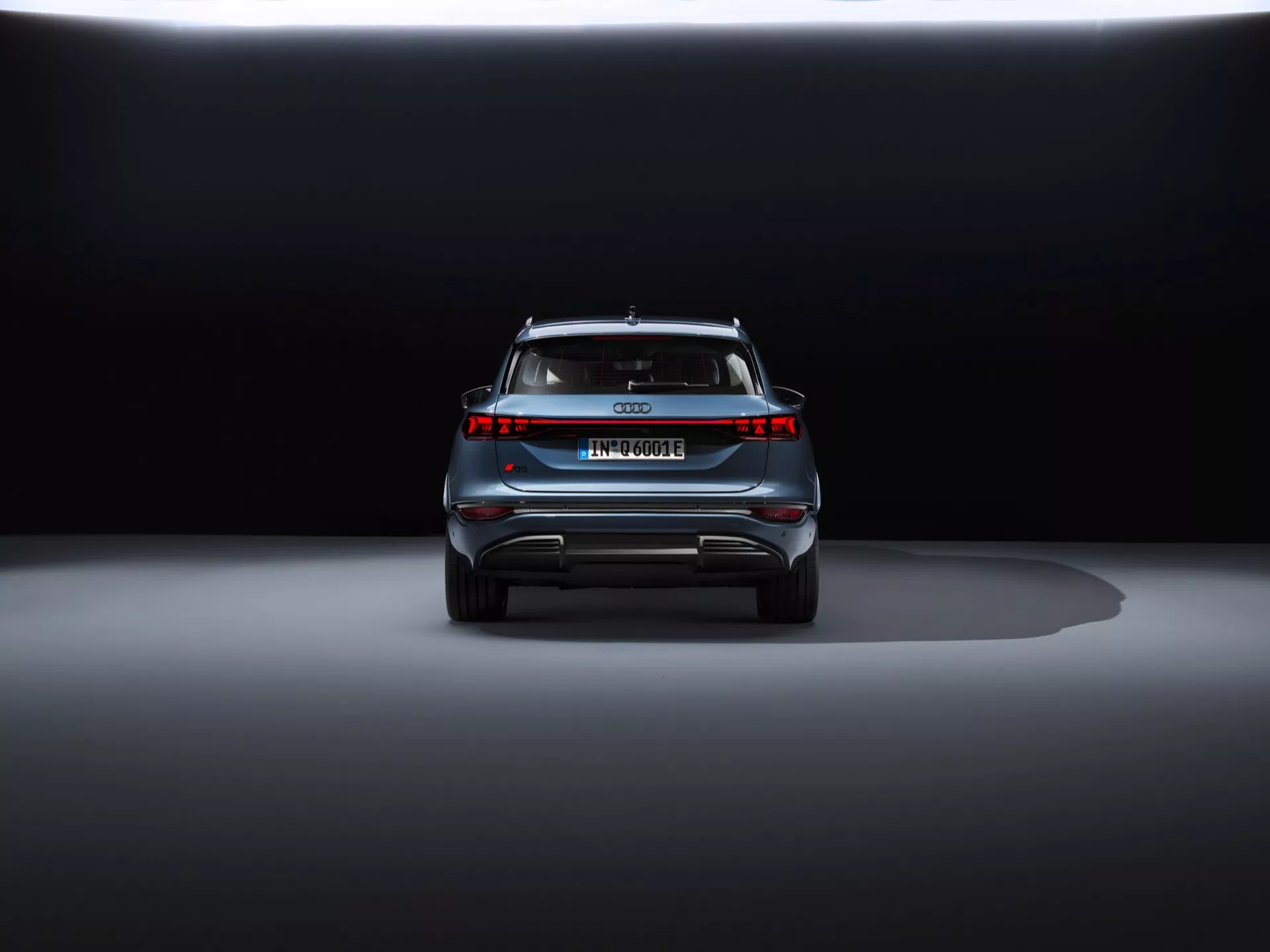 Widok tylu Audi Q6 e-tron 2024 na ciemnym tle, ukazujący elegancki design, centralny emblemat, nazwę modelu, poziomy pasek świetlny, tylny spojler i zderzak w stylu dyfuzora z elementami odblaskowymi.