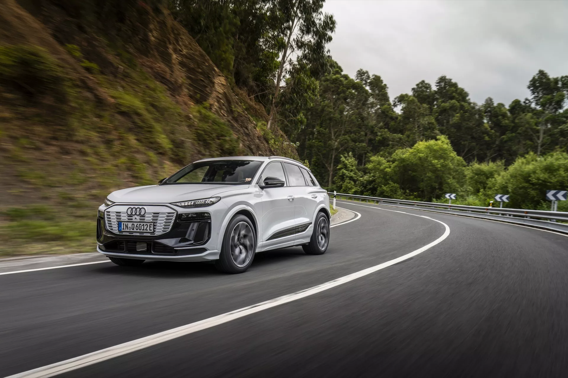 Obraz uchwyca dynamiczny boczny widok nowoczesnego, białego SUV-a, prawdopodobnie modelu Audi Q6 e-tron 2024, jadącego drogą otoczoną zalesionym terenem. Uwaga skupiona jest na przednim grillu, reflektorach, bocznym profilu, kołach i aerodynamice bocznych lusterek pojazdu. Reflektory są eleganckie z technologią LED, co sugeruje zaawansowane oświetlenie. Koła ze stopów lekkich z wieloszprychowym wzorem sugerują wysoką wydajność. Ruch pojazdu na zakręcie podkreśla jego możliwości manewrowe i zaawansowane systemy wspomagania jazdy. Rozmyte tło podkreśla prędkość i skuteczność w nawigacji w trudnych scenariuszach.