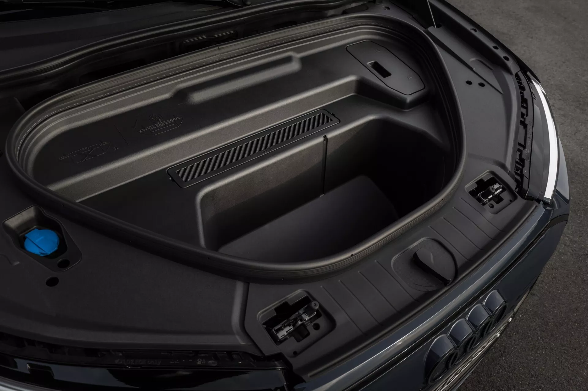 Zbliżenie z wysokiego kąta na tylną przestrzeń bagażnika Audi Q6-e-Tron 2024, ukazujące cechy wnętrza, w tym formowane panele z tworzywa sztucznego dla trwałości, gniazda akcesoriów zasilających po prawej stronie, wbudowany schowek na dole oraz niebieską pokrywkę po lewej stronie, co wskazuje na zaawansowaną technologię samochodową i eleganckie, premium wykończenie.