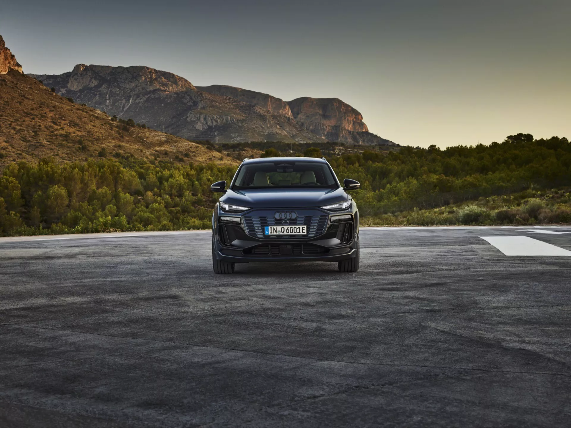 Obraz modelu Audi Q6 e-tron 2024 sfotografowanego pod kątem trzech czwartych z przodu, ukazujący przedni grill pojazdu z emblematem marki, ostre reflektory LED lub laserowe oraz szczegółowy przedni zderzak z wlotami powietrza i możliwymi czujnikami lub kamerami wskazującymi na systemy wspomagania kierowcy. Maskę ozdabiają kontury, a elegancki boczny profil sugeruje aerodynamiczność, z wyższą pozycją SUV-a. Samochód umieszczony jest na zewnątrz na tle naturalnej scenerii, wskazującej na wschód lub zachód słońca.