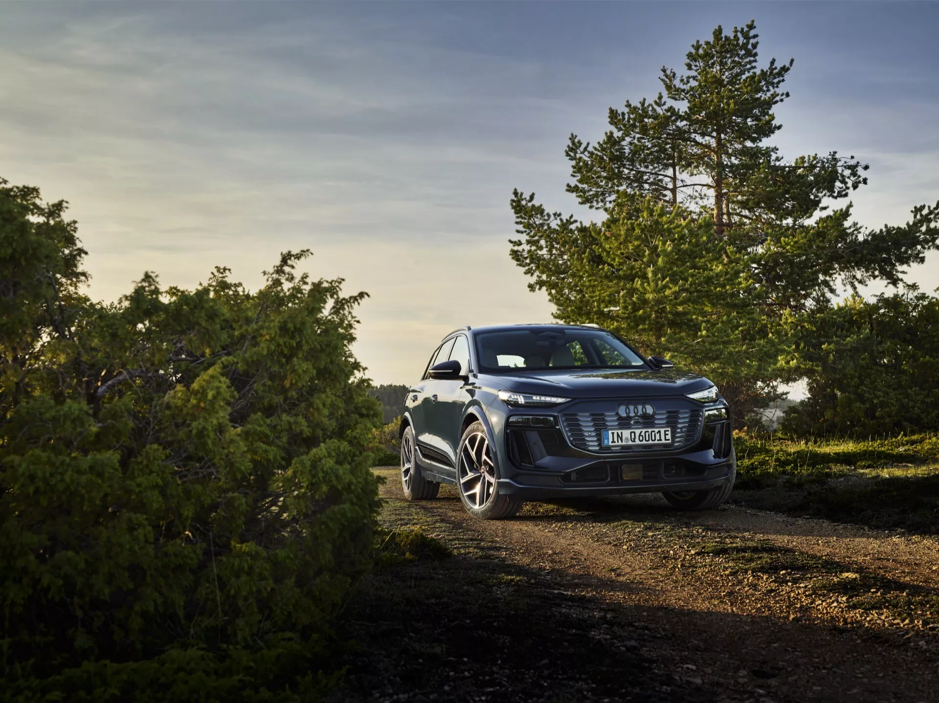Trzy czwarte widoku przedniego Audi Q6 e-tron 2024 zaparkowanego na świeżym powietrzu na drodze szutrowej wśród natury, sfotografowane w ciepłym, złocistym świetle poranka lub późnego popołudnia. Podwyższony kąt ukazuje odważny przedni grill z centralnym emblematem, eleganckie reflektory sugerujące zaawansowane technologie LED lub Matrix LED oraz charakterystyczne sportowe felgi. Zdjęcie zawiera elementy przyrody w tle, wskazując na wszechstronne możliwości samochodu zarówno w środowisku miejskim, jak i na lekkich trasach terenowych.
