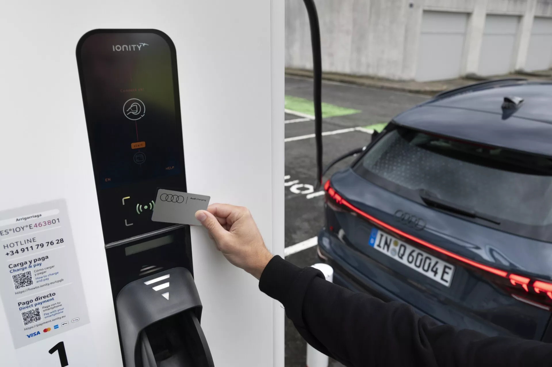 Obraz osoby trzymającej kartę RFID przy czytniku stacji ładowania pojazdów elektrycznych IONITY, skierowany na tylną część nowoczesnego, ciemnego samochodu z poziomymi światłami LED, co sugeruje jego elektryczną naturę. Samochód, prawdopodobnie EV, jest zaparkowany obok ładowarki, co sugeruje, że jest w trakcie ładowania lub przygotowuje się do ładowania.