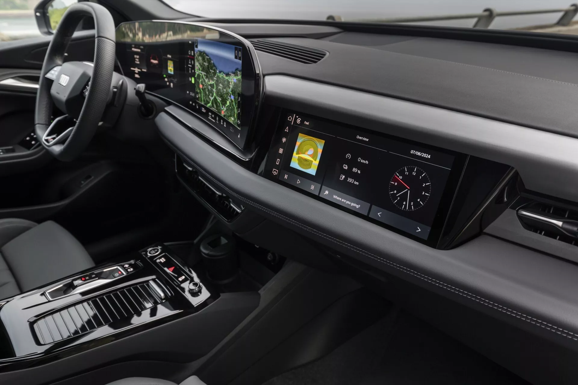 Widok wnętrza Audi Q6 e-tron 2024, skoncentrowany na desce rozdzielczej i konsoli centralnej z lekko podwyższonego kąta. Po lewej stronie widoczna jest kierownica, wskazująca konfigurację do jazdy po lewej stronie, podczas gdy konsola centralna zawiera dźwignię zmiany biegów, różne kontrolki oraz duży wyświetlacz dotykowy. Cyfrowy wyświetlacz przyrządów pokazuje metryki jazdy. Stylowa deska rozdzielcza rozciąga się poziomo z otworami wentylacyjnymi i innym elementem wyświetlacza po prawej stronie. Zaawansowana technologia jest widoczna dzięki centralnemu ekranowi dotykowemu, który pokazuje informacje o nawigacji i multimediach, wyświetlaczowi w polu widzenia widocznemu nad kierownicą oraz dotykowym przyciskom do sterowania klimatyzacją. Projekt podkreśla minimalizm i materiały premium, ze skórzanymi przeszyciami i metalowymi akcentami, które podkreślają luksusowy charakter wnętrza.