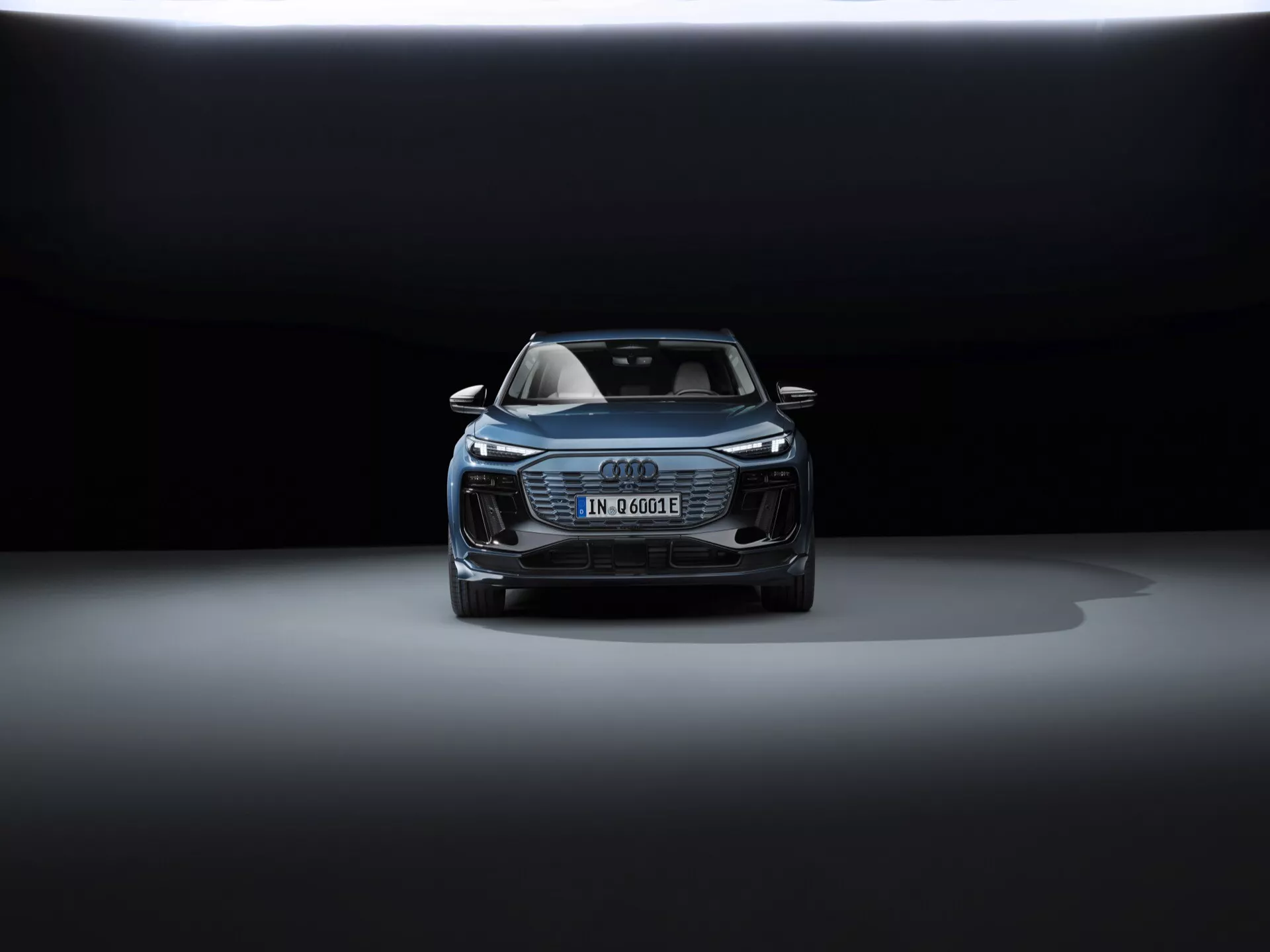 Widok pod kątem na przód Audi Q6 e-tron 2024, pokazujący maskę, grill, część dachu i przedni design. Eleganckie reflektory prawdopodobnie wyposażone są w technologię LED. Niemiecka tablica rejestracyjna sugeruje elektryczny lub hybrydowy charakter pojazdu. Charakterystyczny grill Audi Singleframe, dodatkowe wloty i potencjalne czujniki bezpieczeństwa, podkreślają jego zaawansowane technologie asystujące kierowcy. Dramatyczne oświetlenie z ciemnym tłem i oświetleniem od góry podkreśla luksusowy i wyrafinowany design samochodu.
