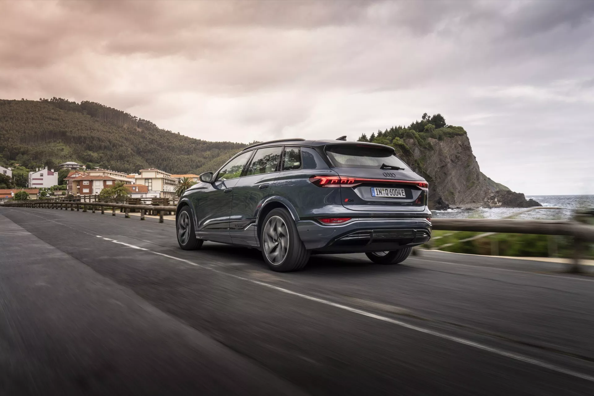 Widok Audi Q6 e-tron 2024 z tyłu pod kątem trzech czwartych w ruchu na przybrzeżnej drodze, prezentujący charakterystyczne tylne światła LED, ciemny metaliczny kolor, aluminiowe felgi i praktyczną tylną wycieraczkę. Rozmyte tło wskazuje na nadmorskie otoczenie, odzwierciedlając estetykę i zaawansowanie technologiczne nowoczesnego SUV-a.